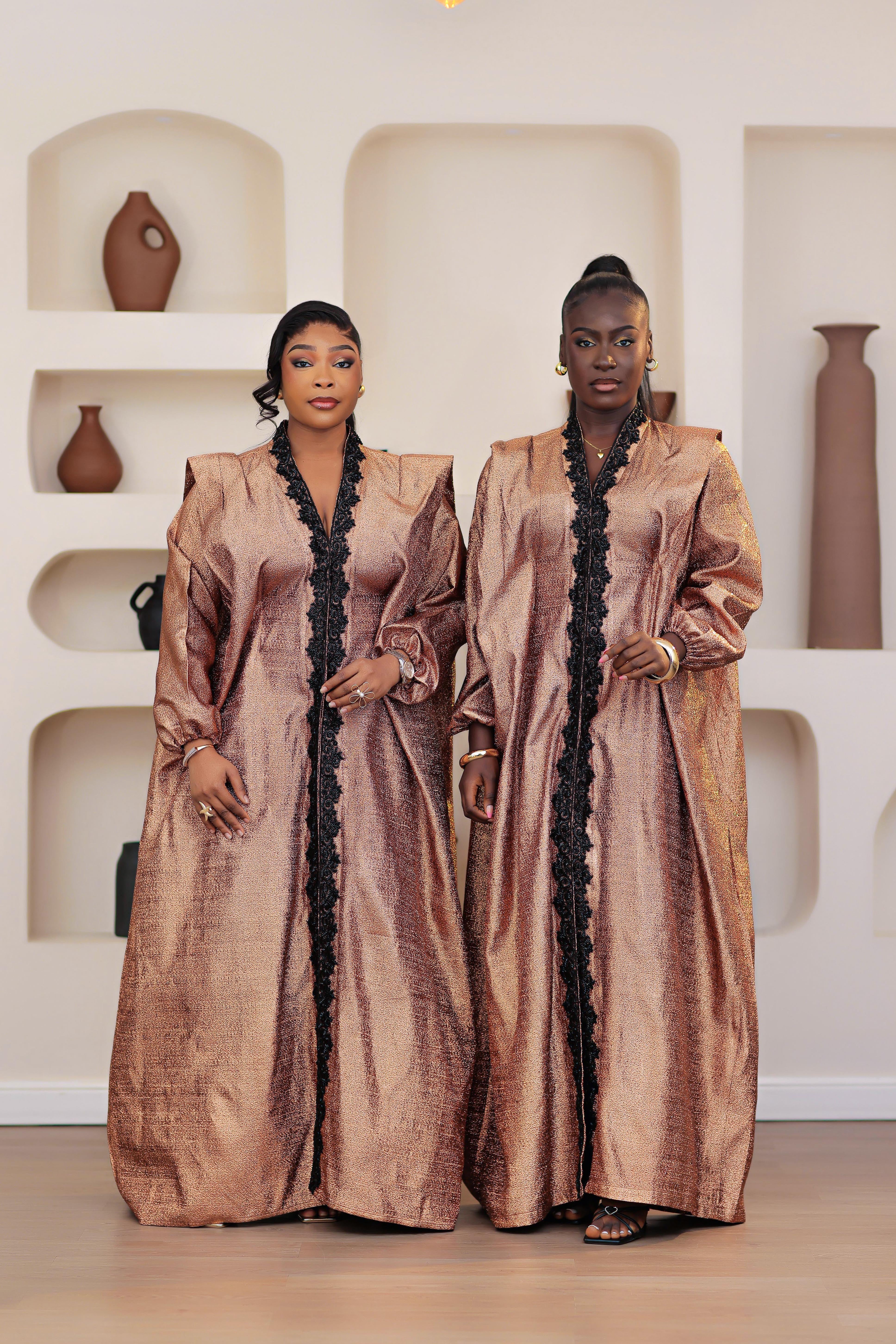 two women in a long, long sleeved African brocade boubou dress with embellished black stone trimmings standing in a room with shelves displaying vases.