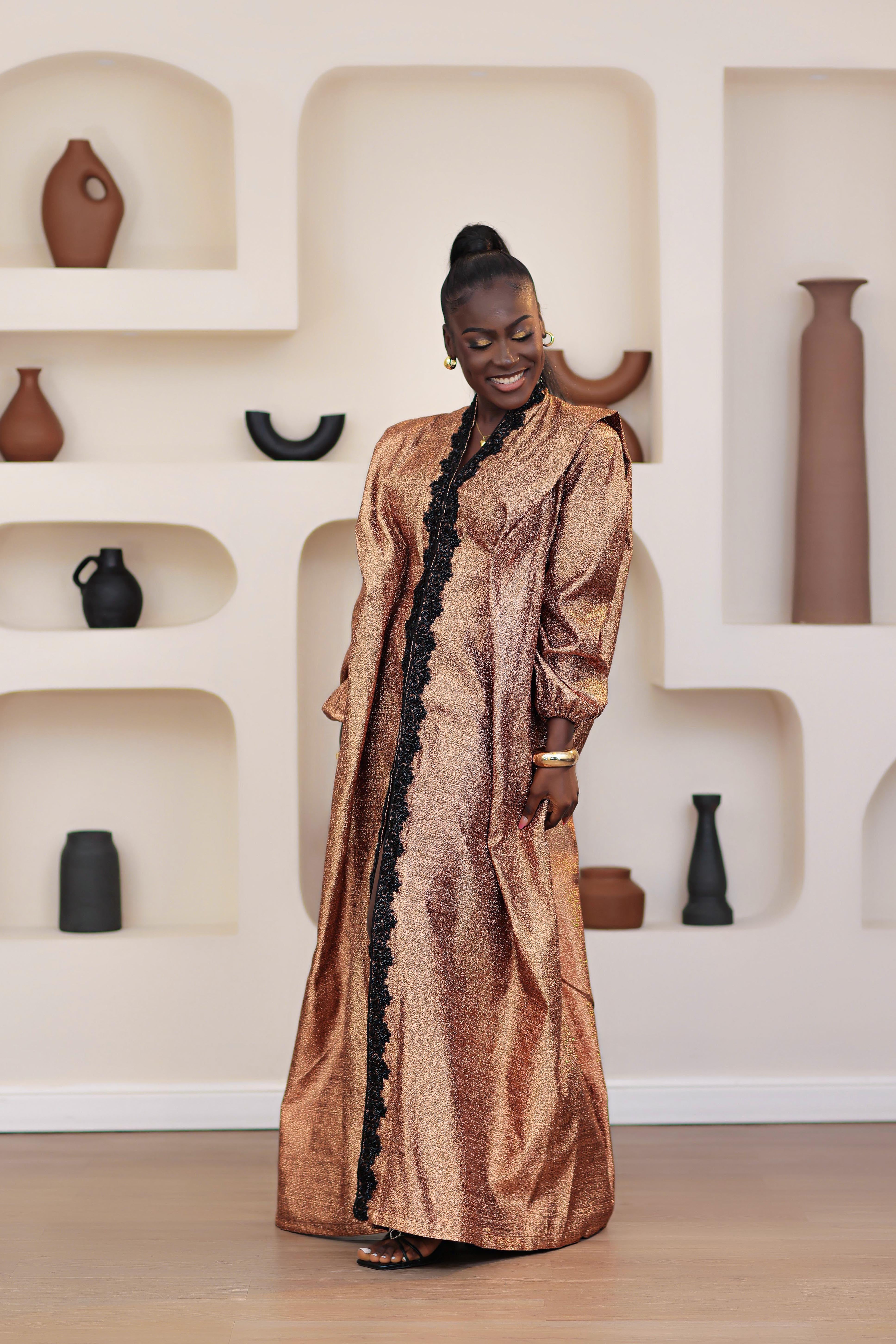 Woman in a long, African brocade boubou dress with embellished black stone trimmings standing in a room with shelves displaying vases.