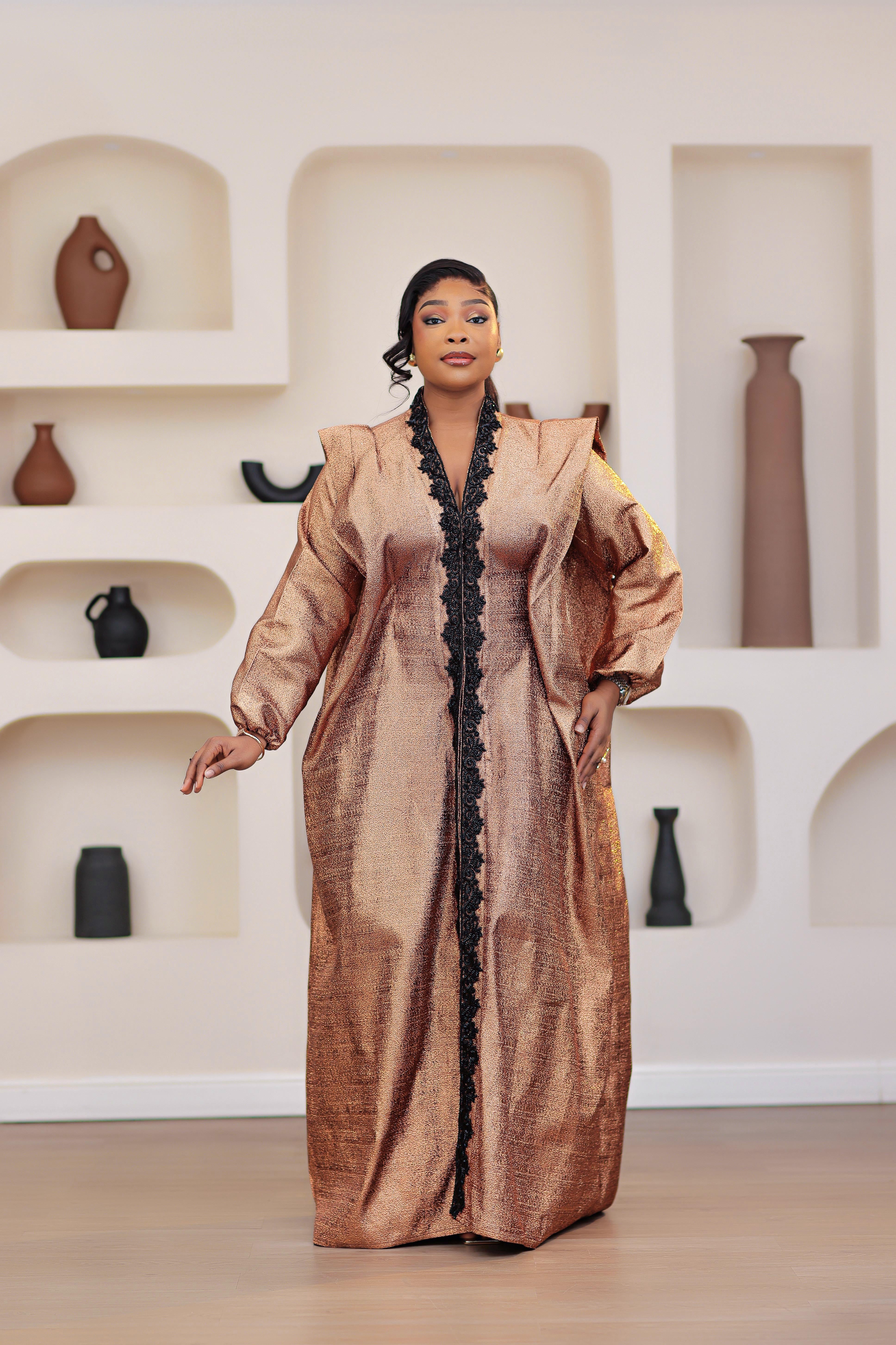 Woman in a long, African brocade boubou dress with embellished black stone trimmings standing in a room with shelves displaying vases.