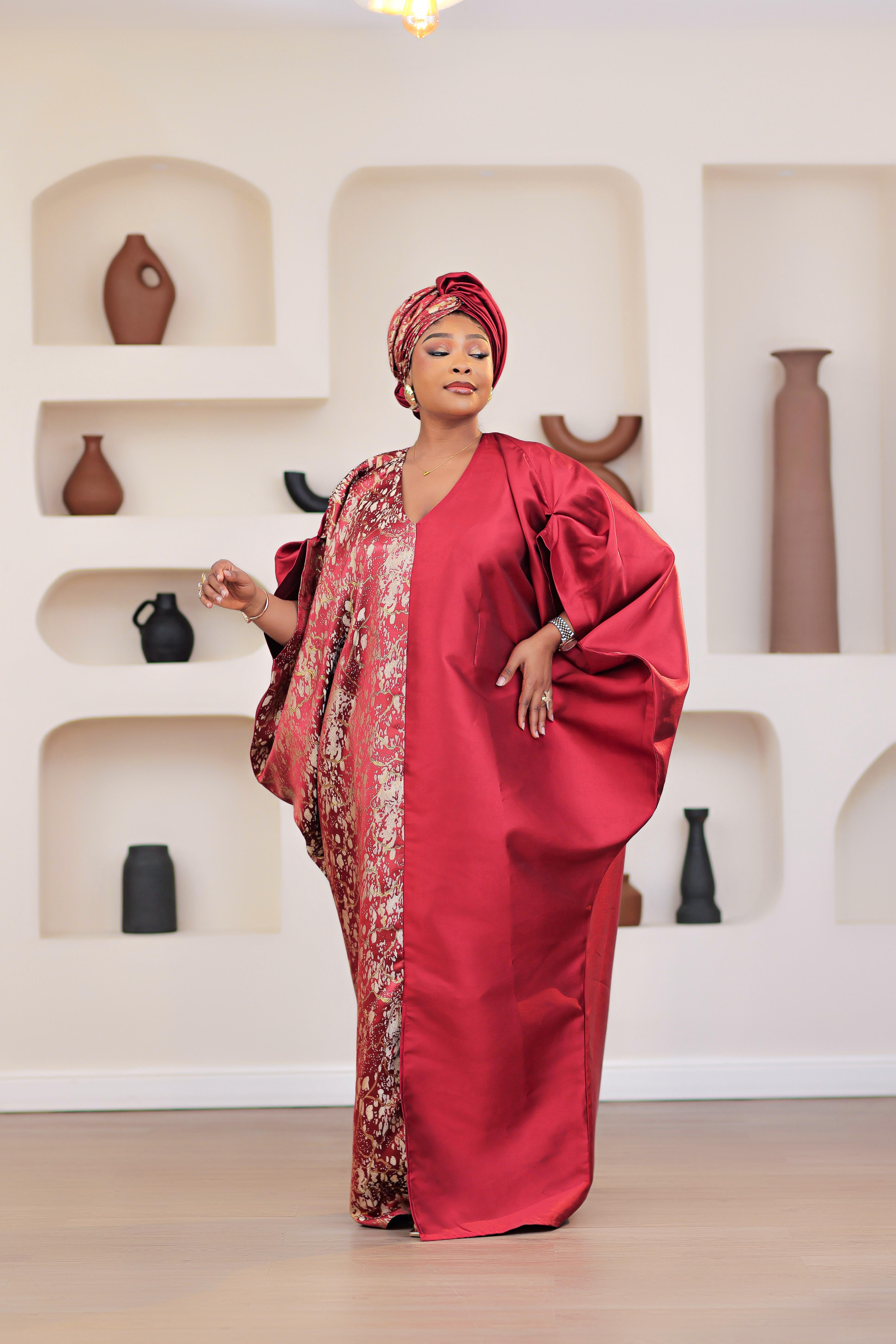 Woman in a red African Brocade Boubou traditional outfit with headwrap standing in front of a decorative wall with shelves.
