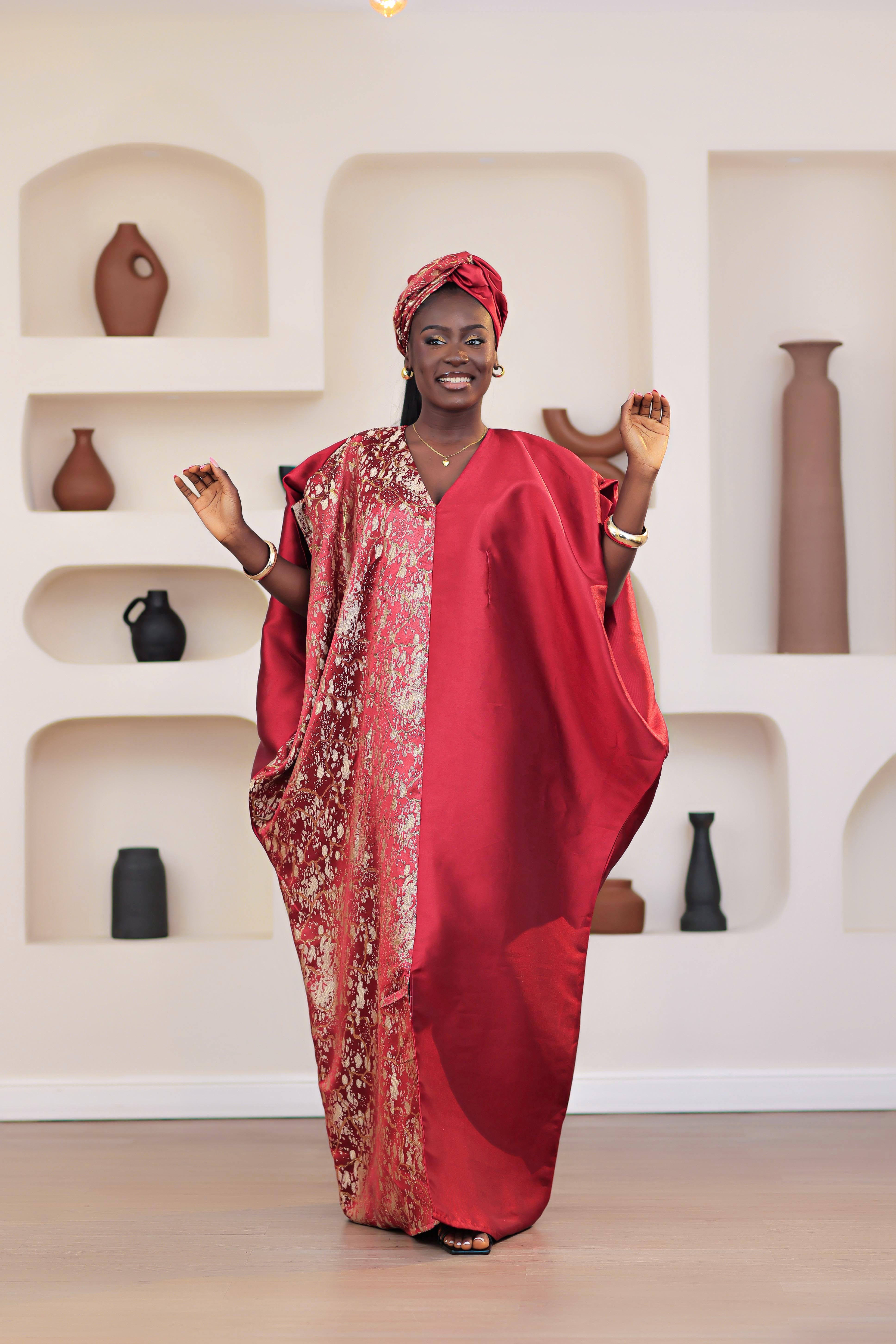 Woman in a red African Brocade Boubou traditional outfit with headwrap standing in front of a decorative wall with shelves.