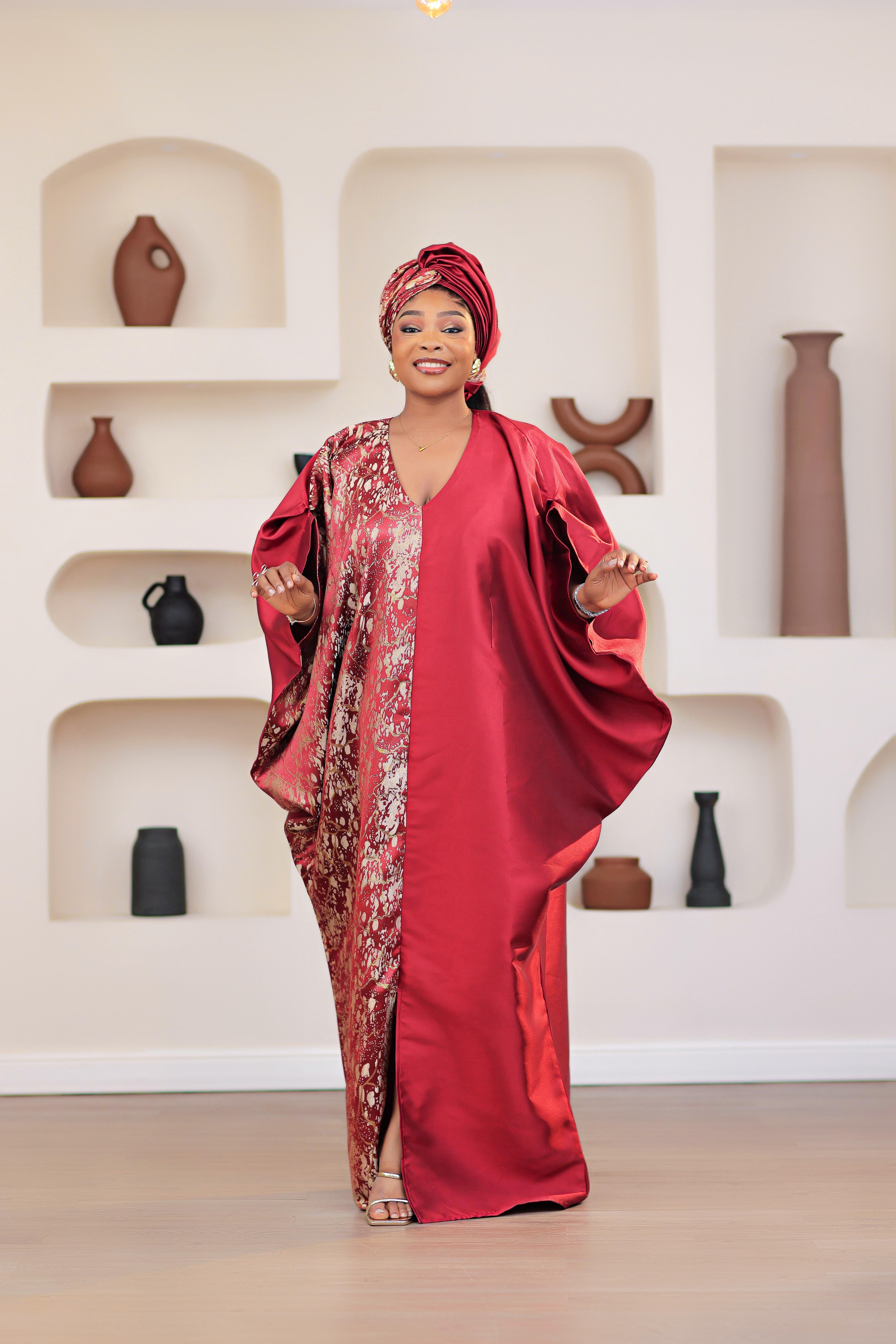Woman in a red African Brocade Boubou traditional outfit with headwrap standing in front of a decorative wall with shelves.