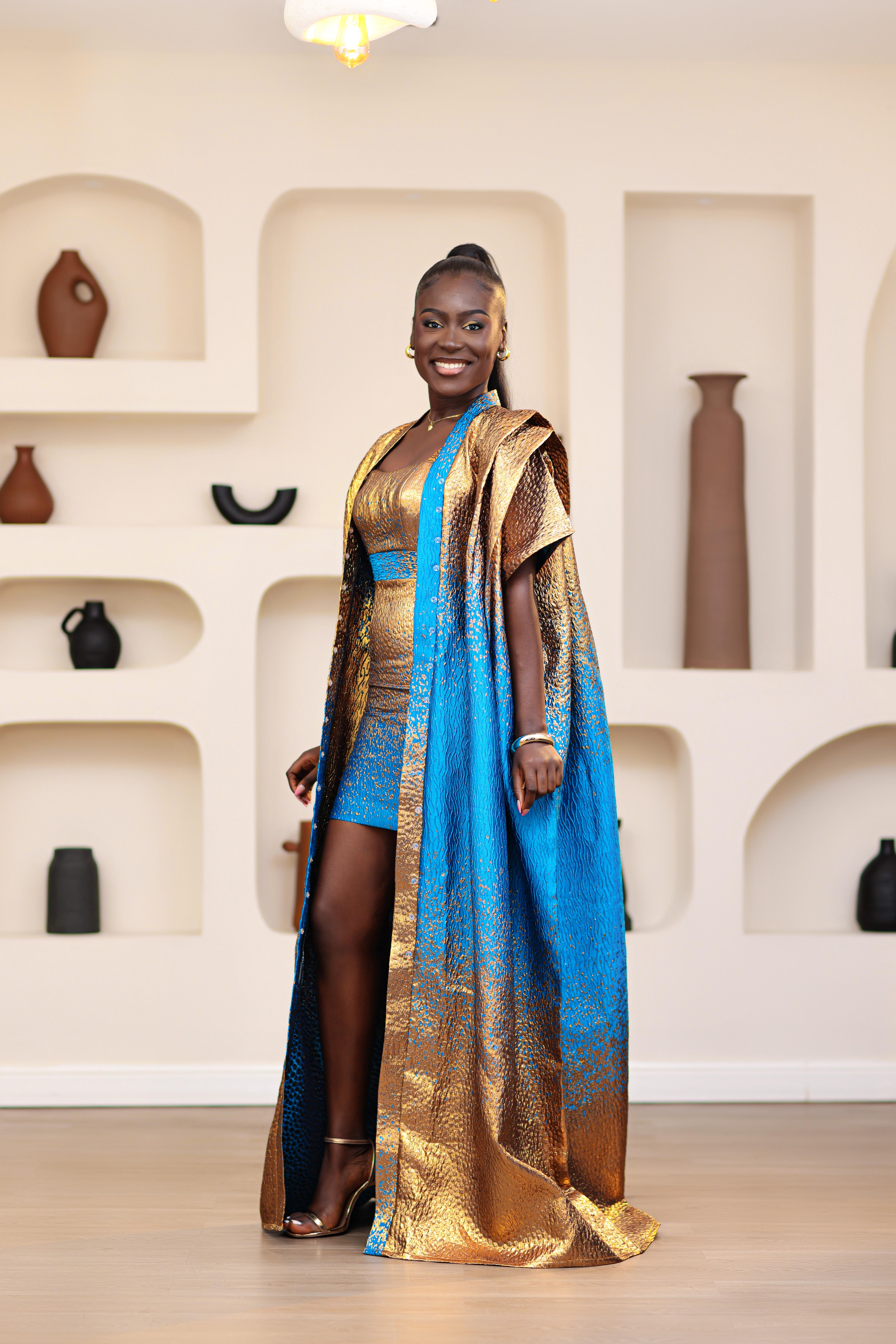 Woman in a blue and gold African traditional Brocade boubou and mini dress outfit set standing in a room with shelves displaying vases.
