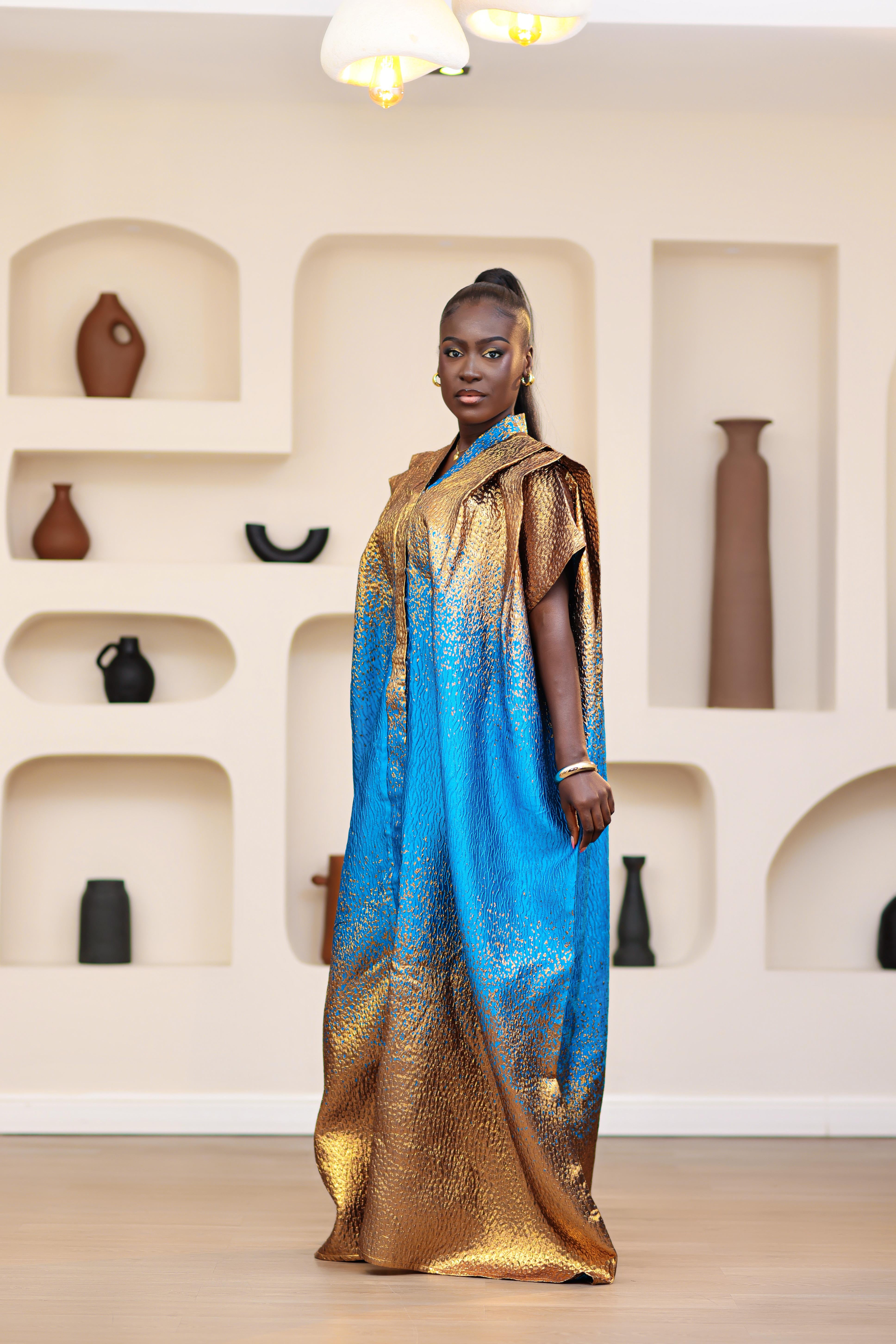 Woman in a blue and gold African traditional Brocade boubou and mini dress outfit set standing in a room with shelves displaying vases.