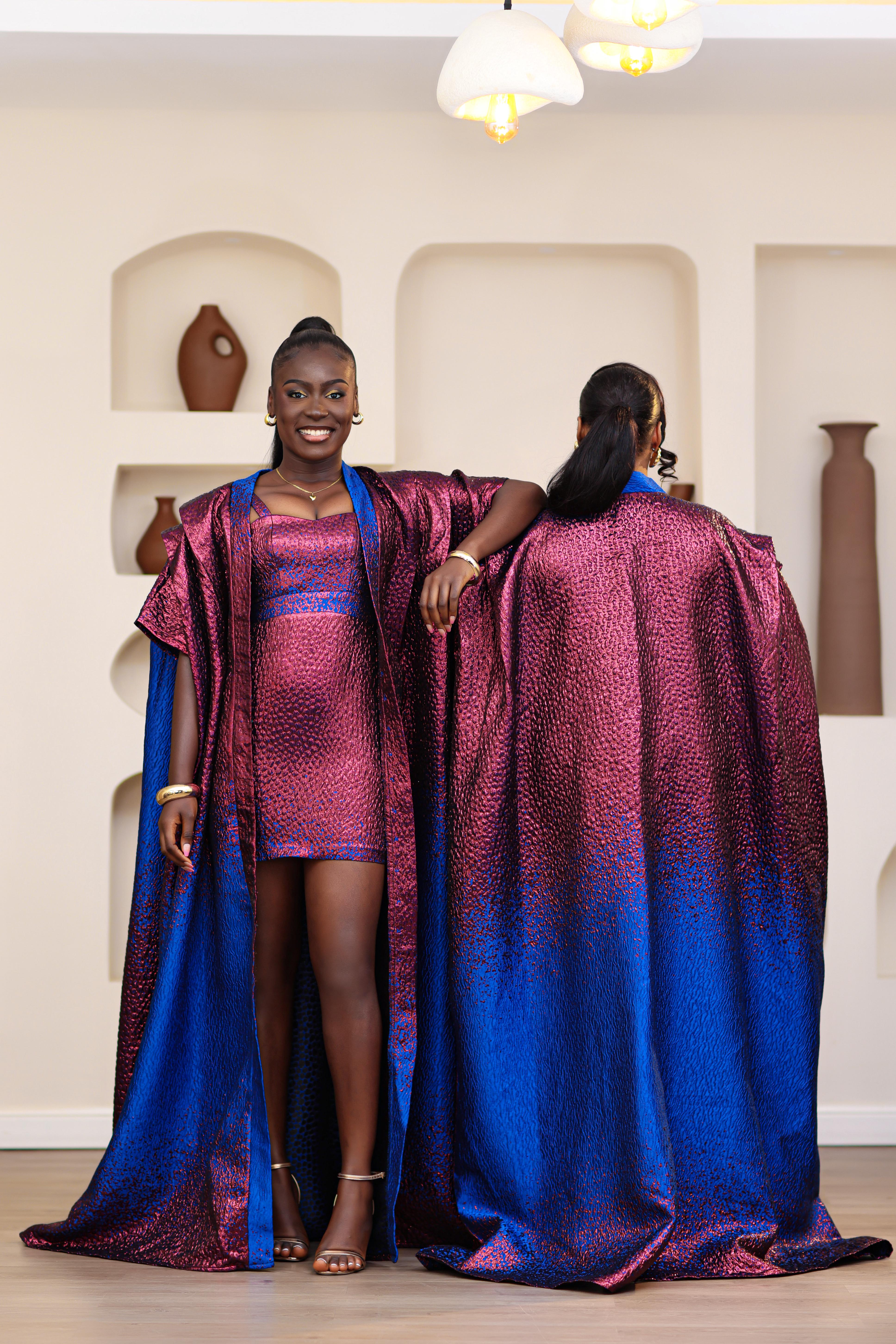 Women in an African brocade blue and pink mini dress with a matching long boubou kimono standing in a room with shelves displaying vases.