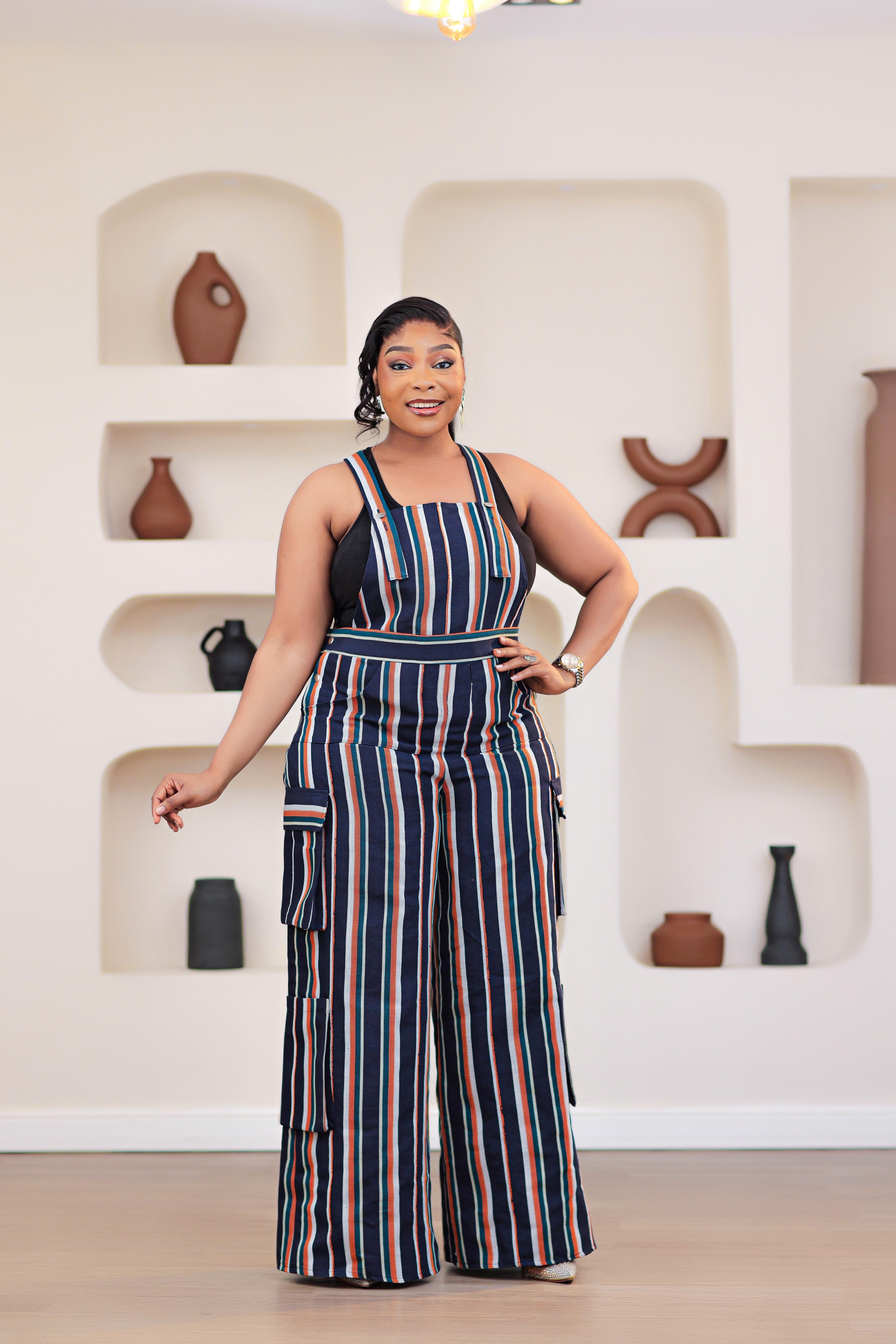 Woman wearing a striped African aso-oke dungarees jumpsuit standing in front of a decorative wall with shelves.
