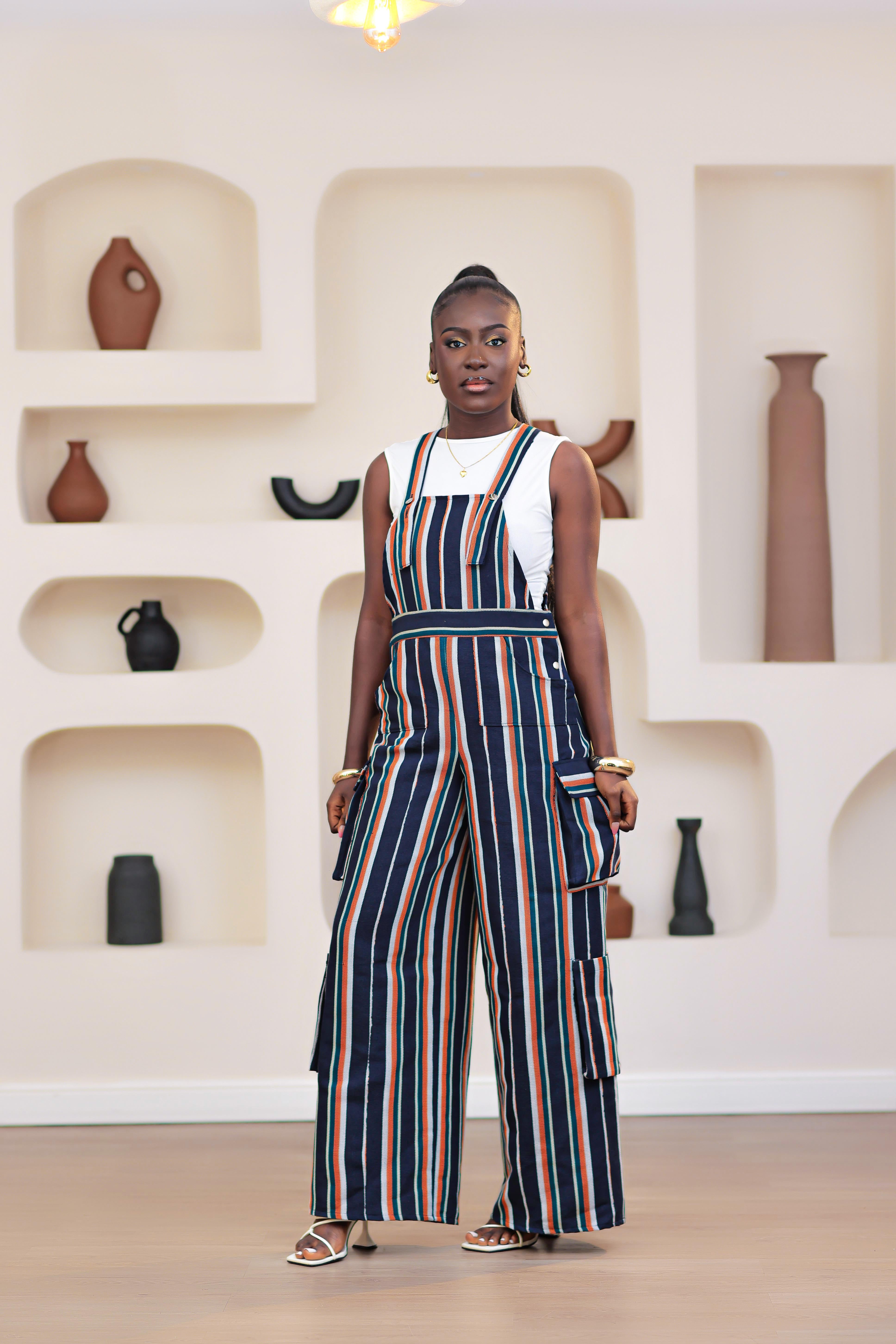 Woman wearing a striped African Aso-oke dungarees jumpsuit standing in front of a wall with shelves displaying vases.