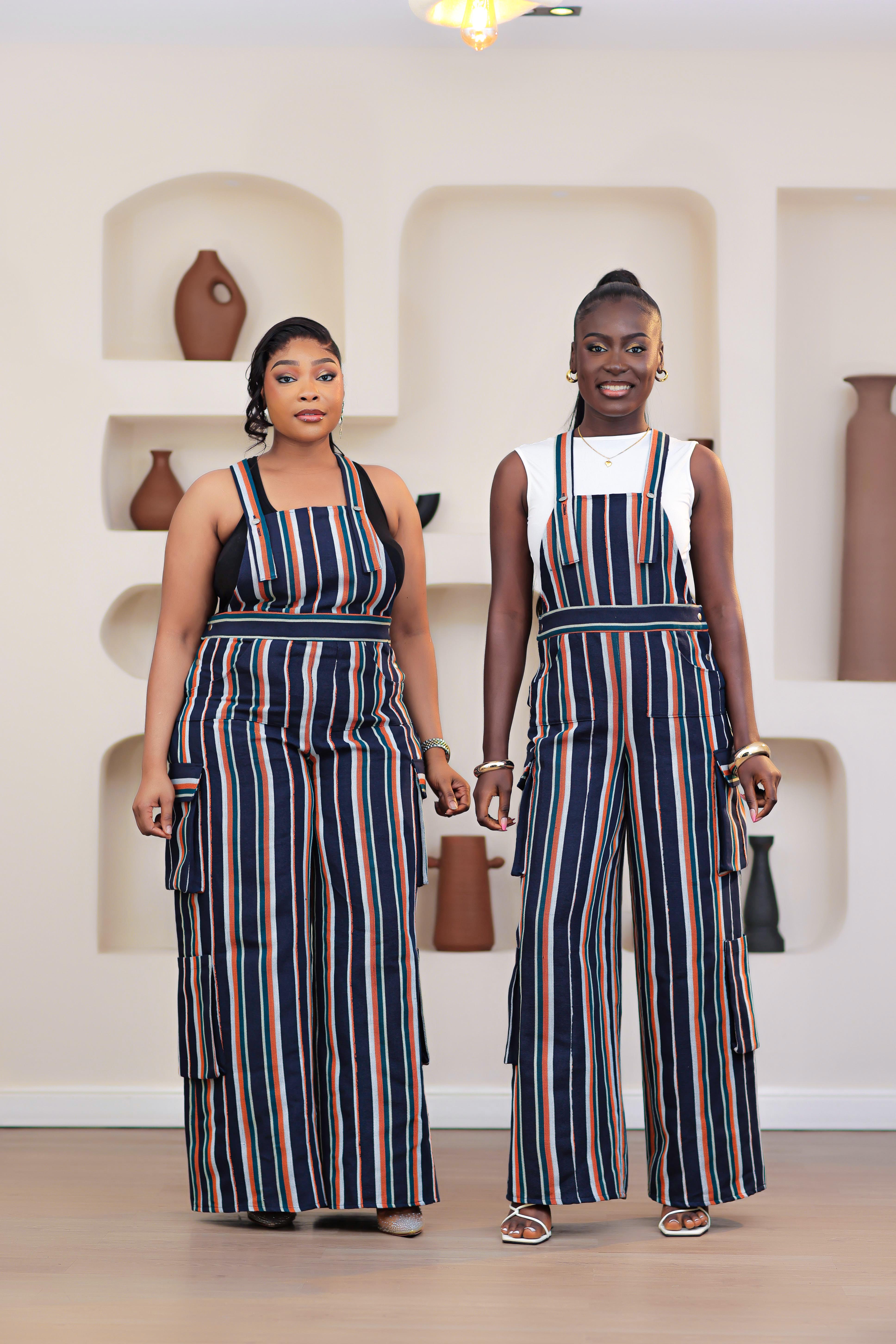 Two women wearing matching striped African Aso-oke dungarees jumpsuit outfits standing in a room with shelves.