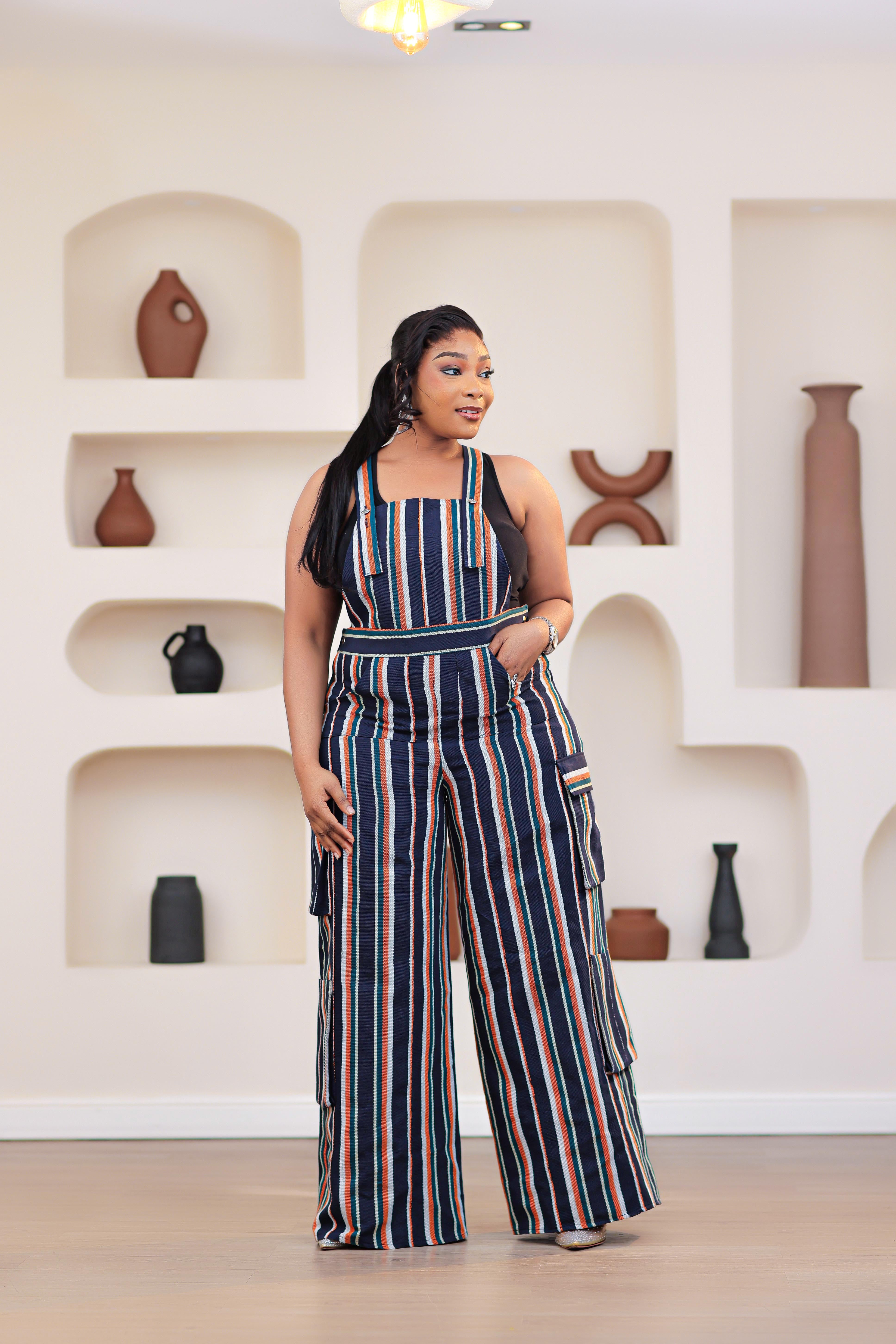 Woman wearing a striped NIKI KAY Aso-oke dungarees jumpsuit standing in a room with shelves and vases.