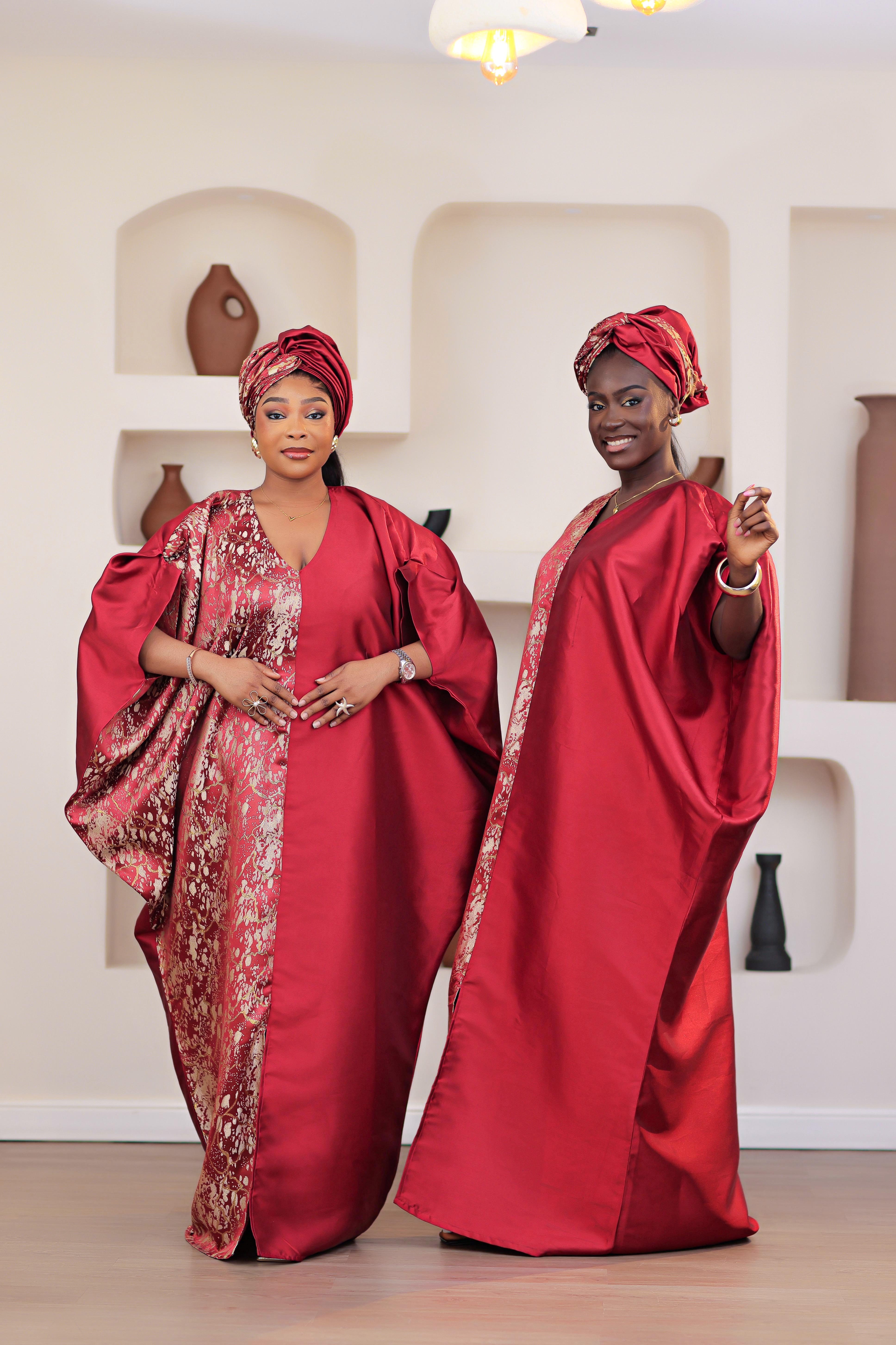 Woman in a red African Brocade Boubou traditional outfit with headwrap standing in front of a decorative wall with shelves.