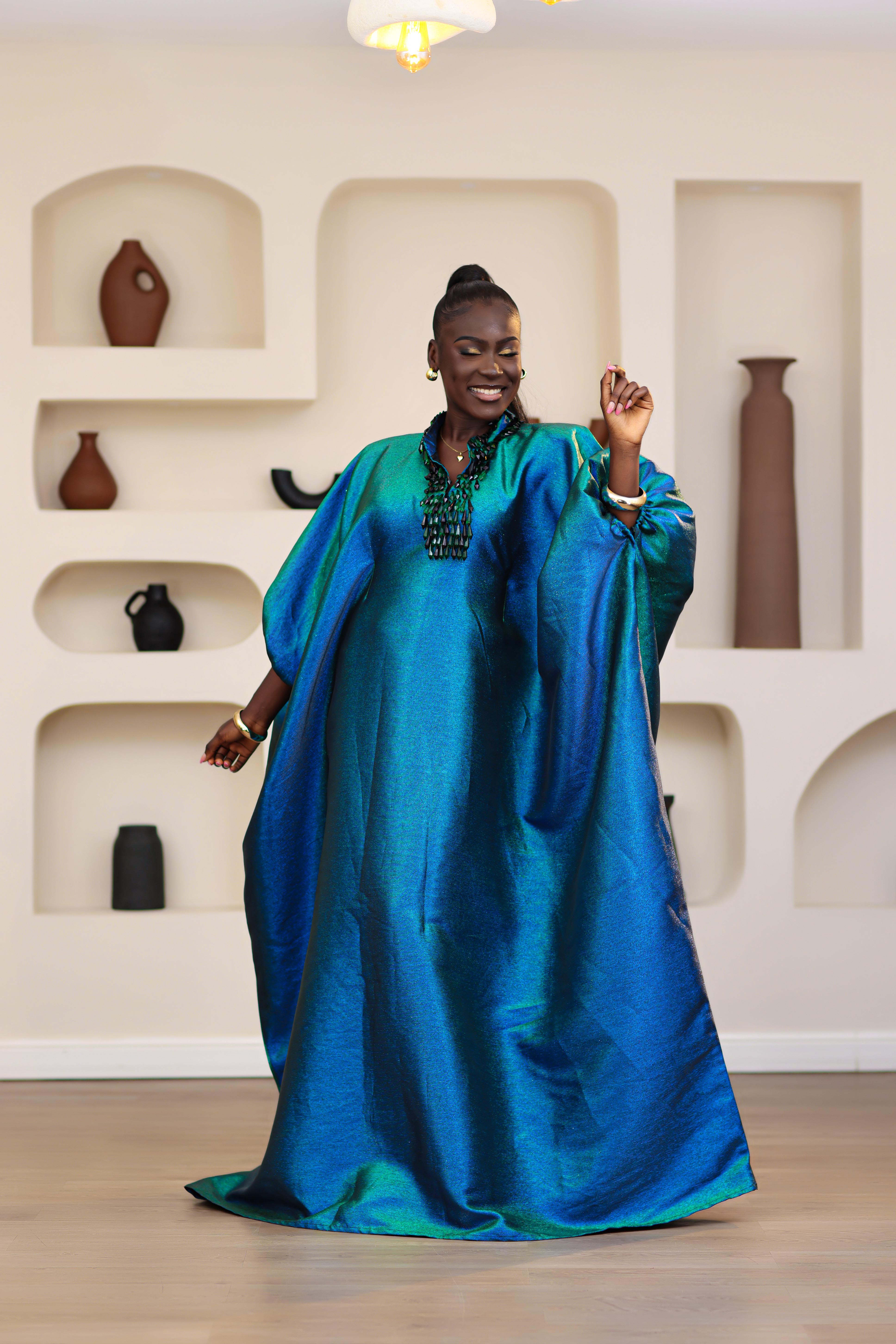 Woman wearing a blue and green NIKI KAY Iridescent brocade boubou dress in a room with decorative vases on shelves.