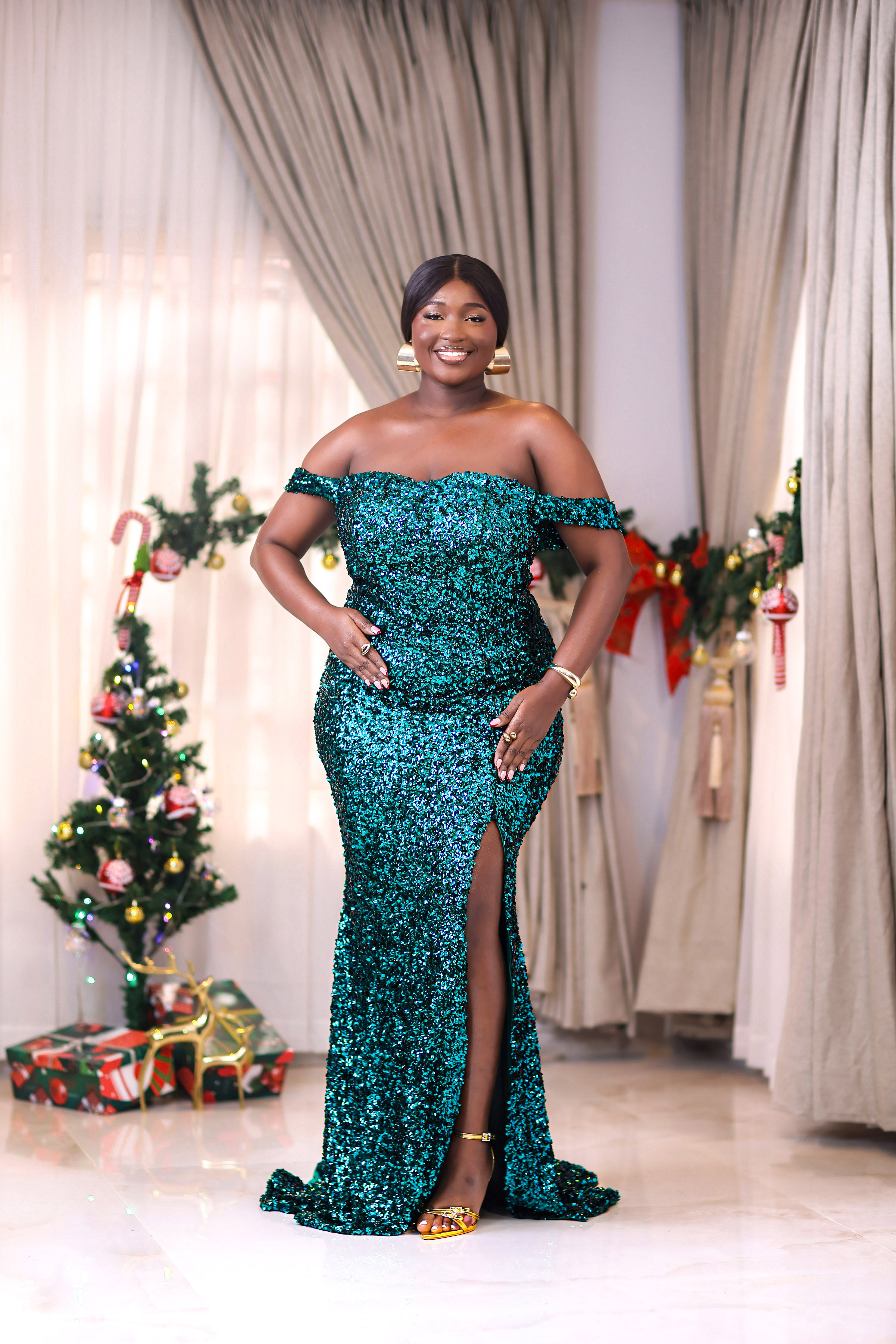 Woman in a sparkling sequin green off-shoulder long slit dress gown standing in a warmly lit indoor setting.