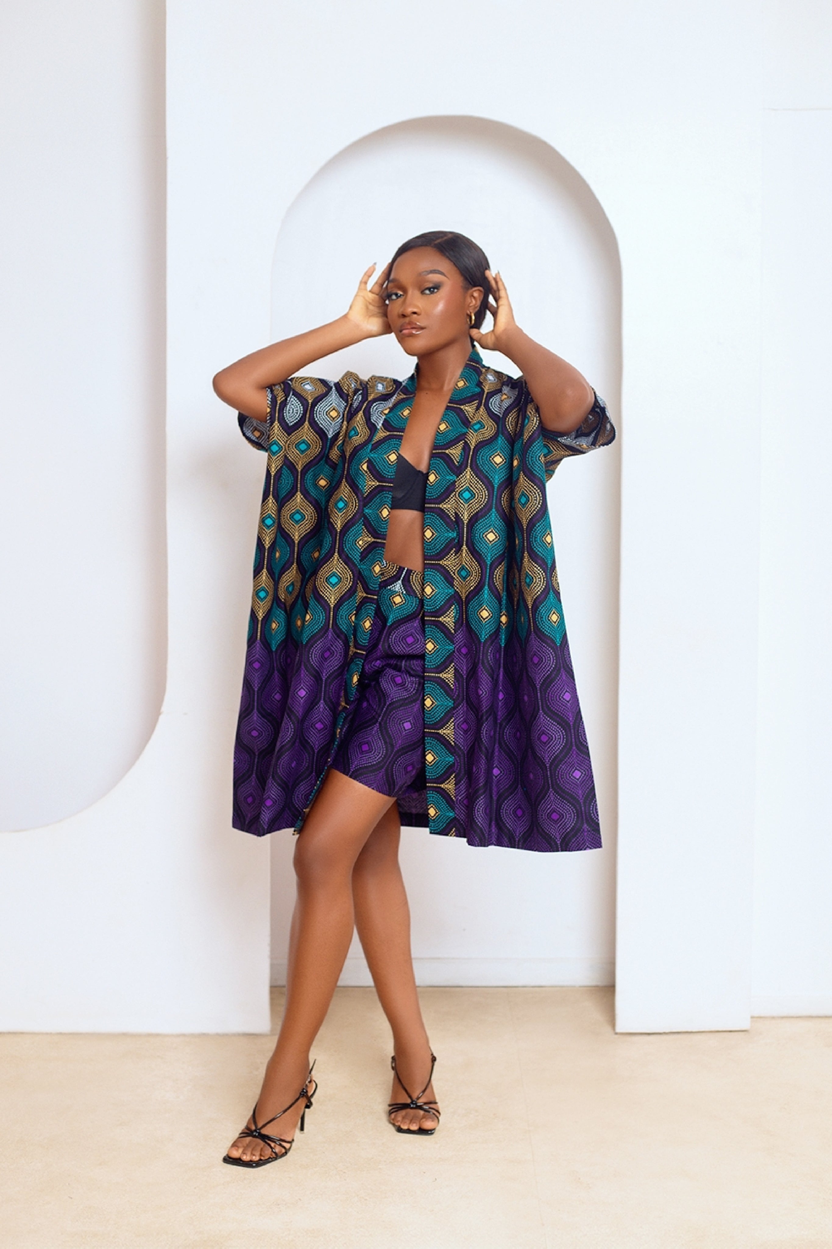 A model wearing a multicolored African print kimono and matching shorts with side pockets. The outfit features a mix of purple, green, and other colors in a geometric pattern.