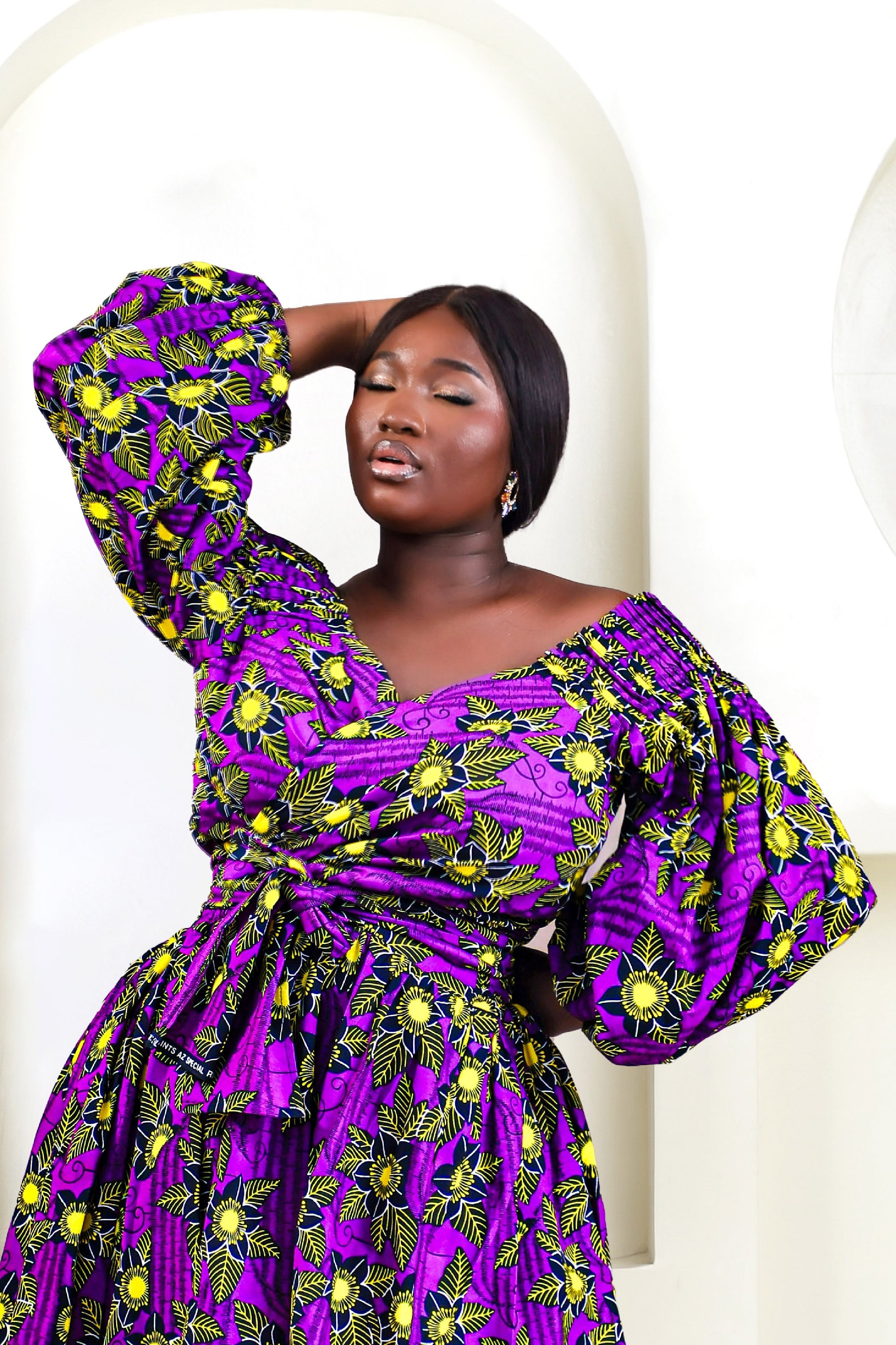 A woman wearing a bold African print Ankara purple and yellow African print wrap top with bubble sleeves and a off-the-shoulder neckline.