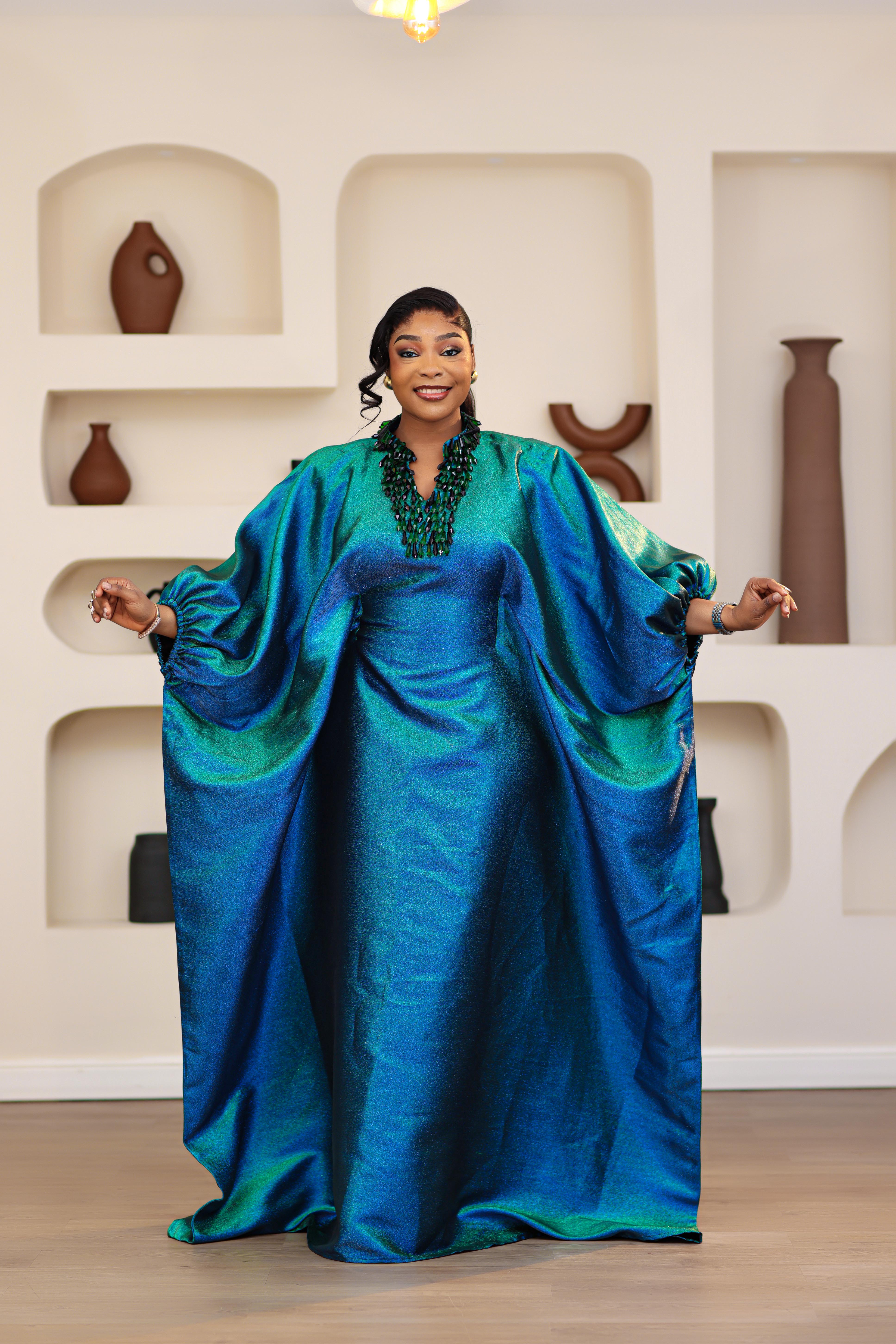 Woman wearing a blue and green NIKI KAY Iridescent brocade boubou dress in a room with decorative vases on shelves.
