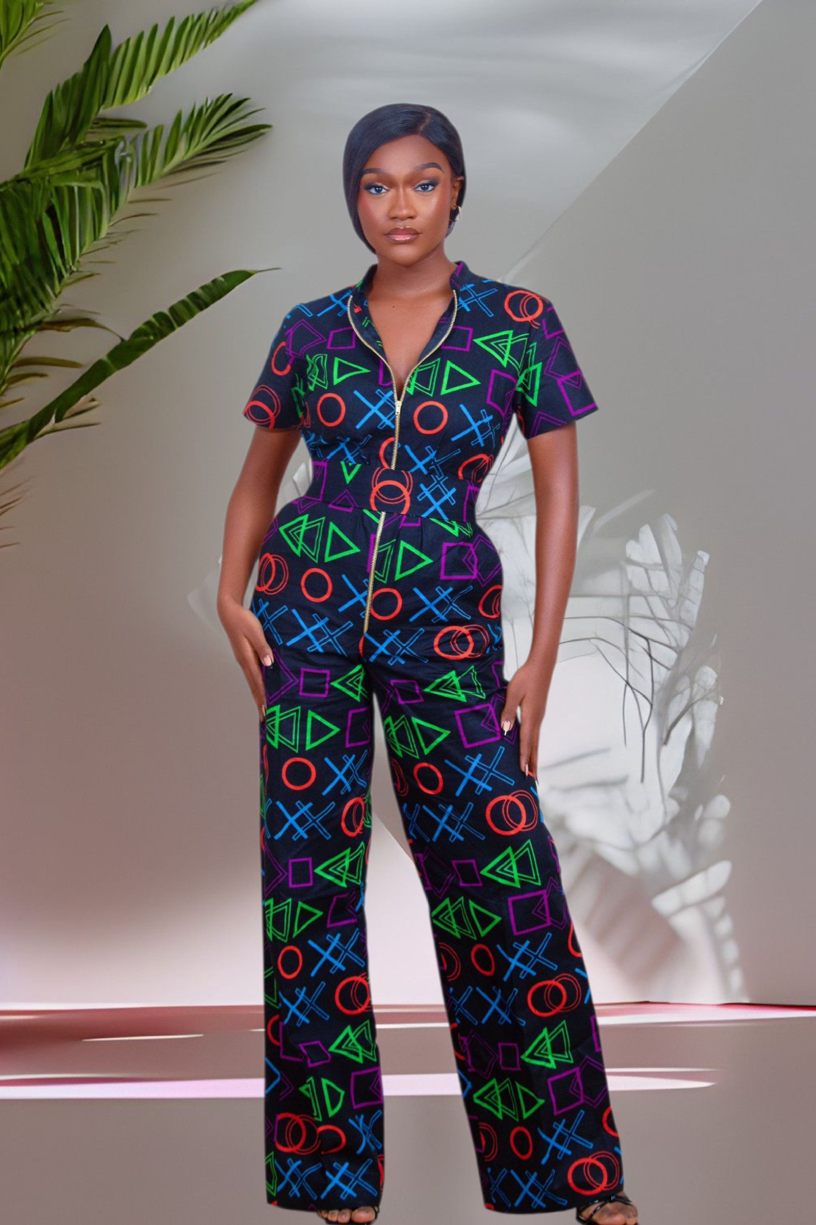 Woman wearing a colorful geometric patterned multicolored jumpsuit standing indoors with a plant in the background.