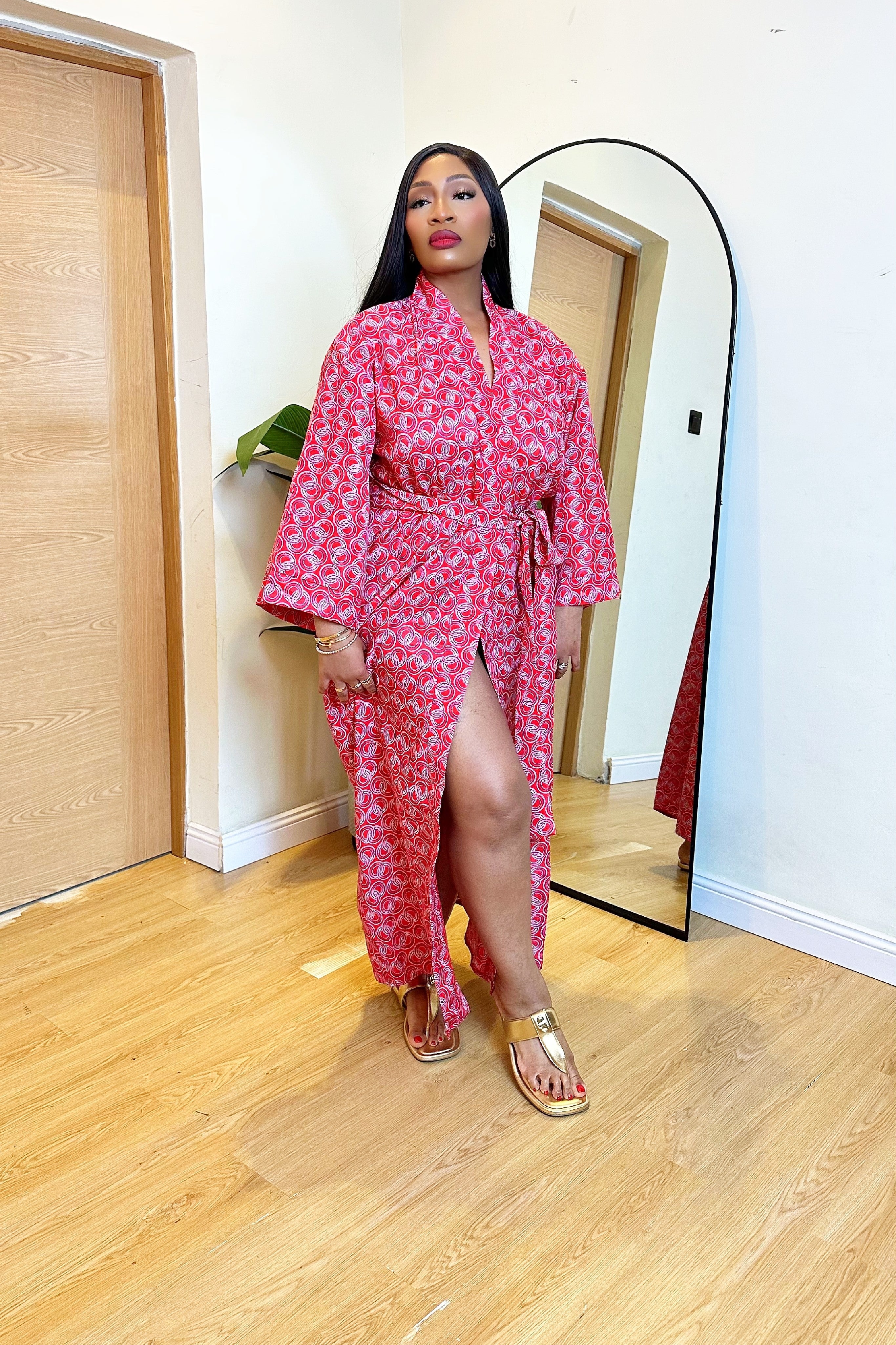 Woman in a colorful red pink African print robe dress standing in front of a mirror in a room with wooden flooring.