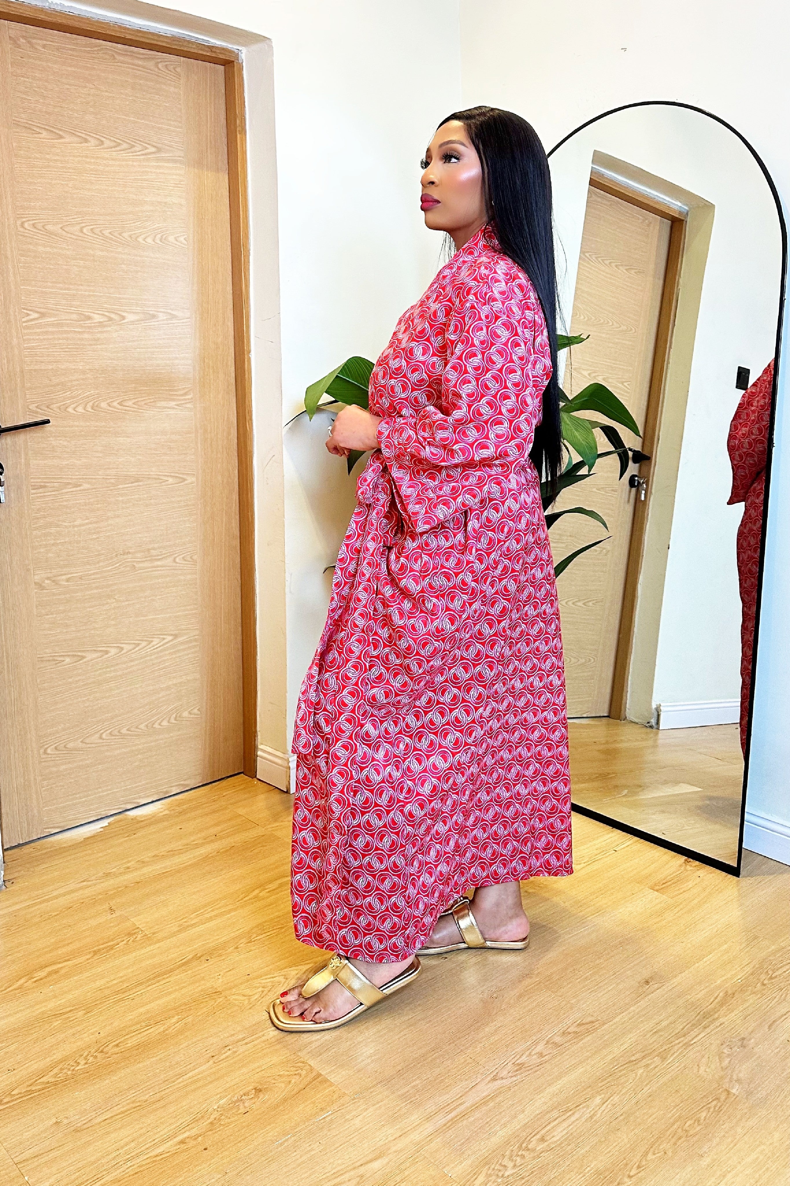 Woman in a colorful red pink African print robe dress standing in front of a mirror in a room with wooden flooring.