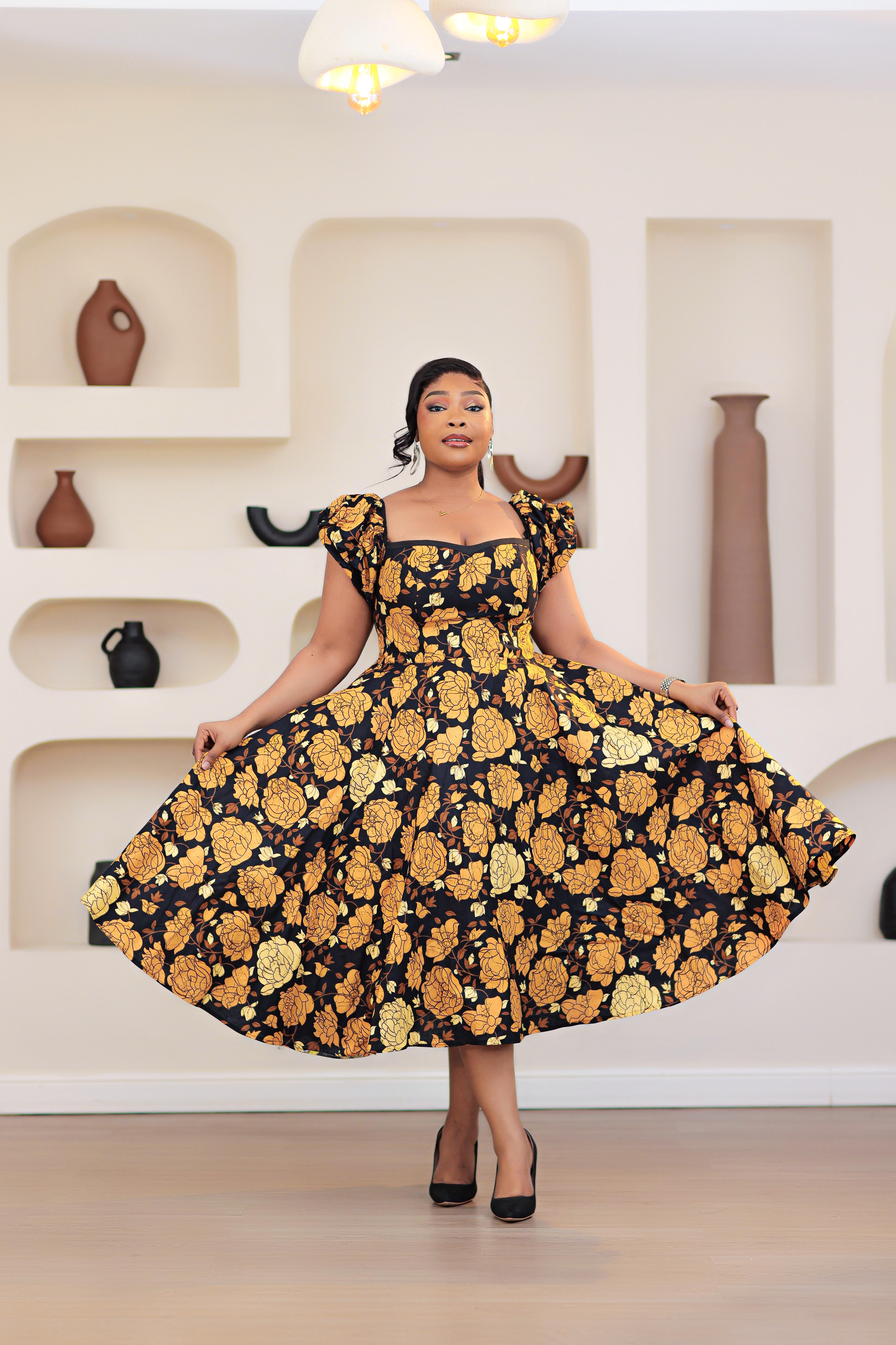 Woman wearing an African print Ankara gold, brown, black, yellow short sleeve floral dress in a room with shelves and vases.