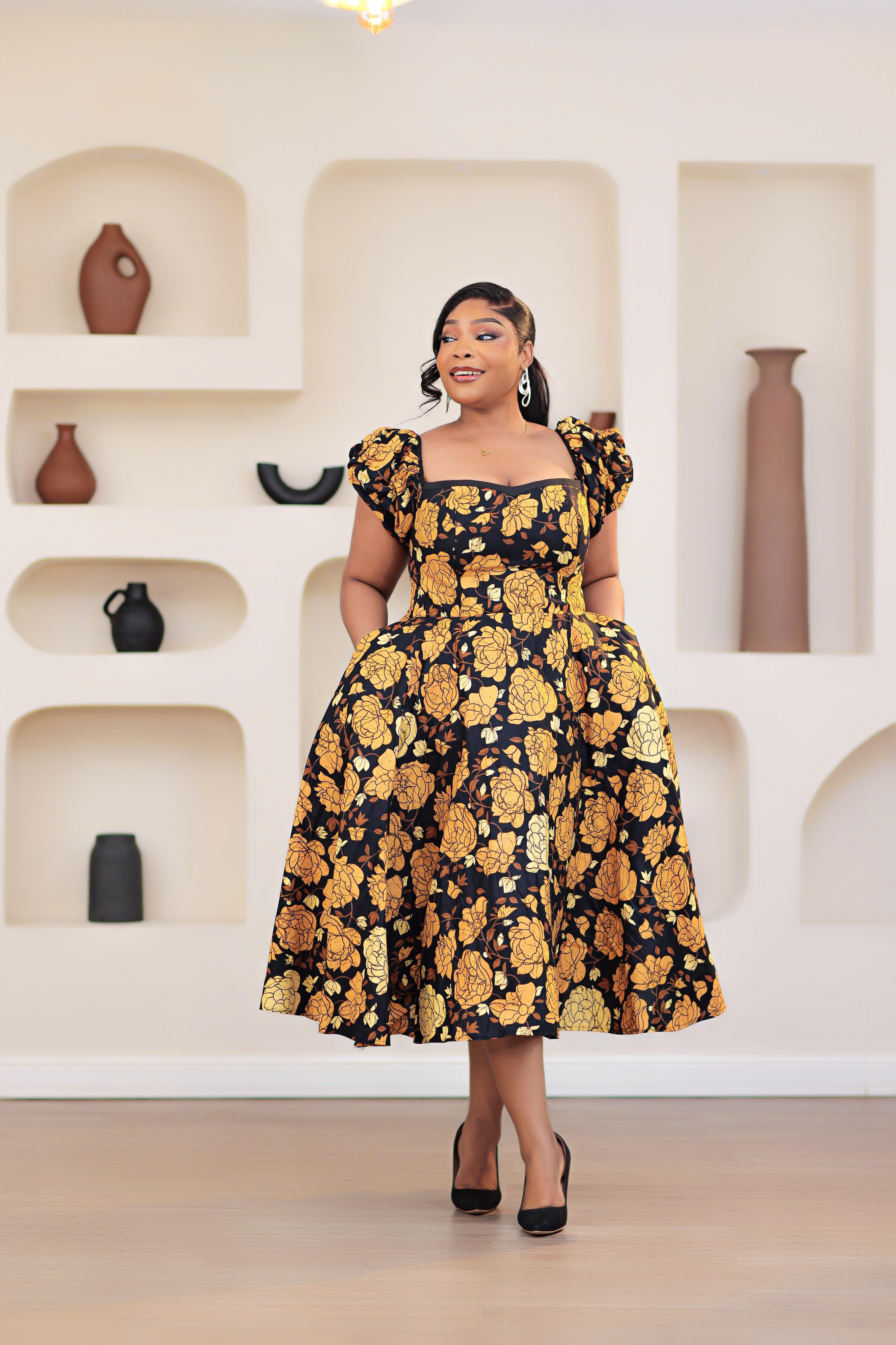Woman wearing an African print Ankara gold, brown, black, yellow short sleeve floral dress in a room with shelves and vases.