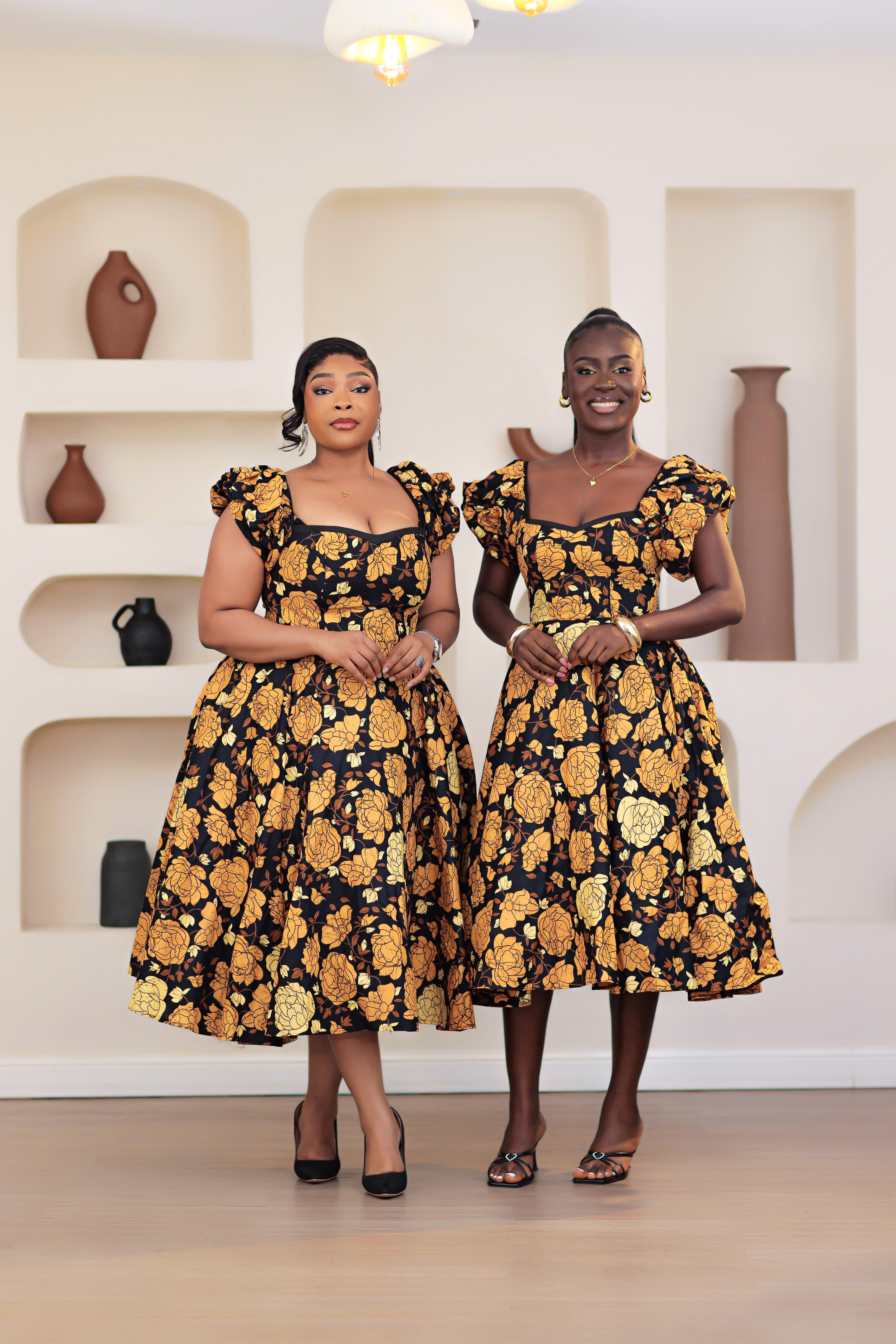 Two women wearing African print Ankara gold, brown, black, yellow floral short sleeve dress in a room with shelves and vases.
