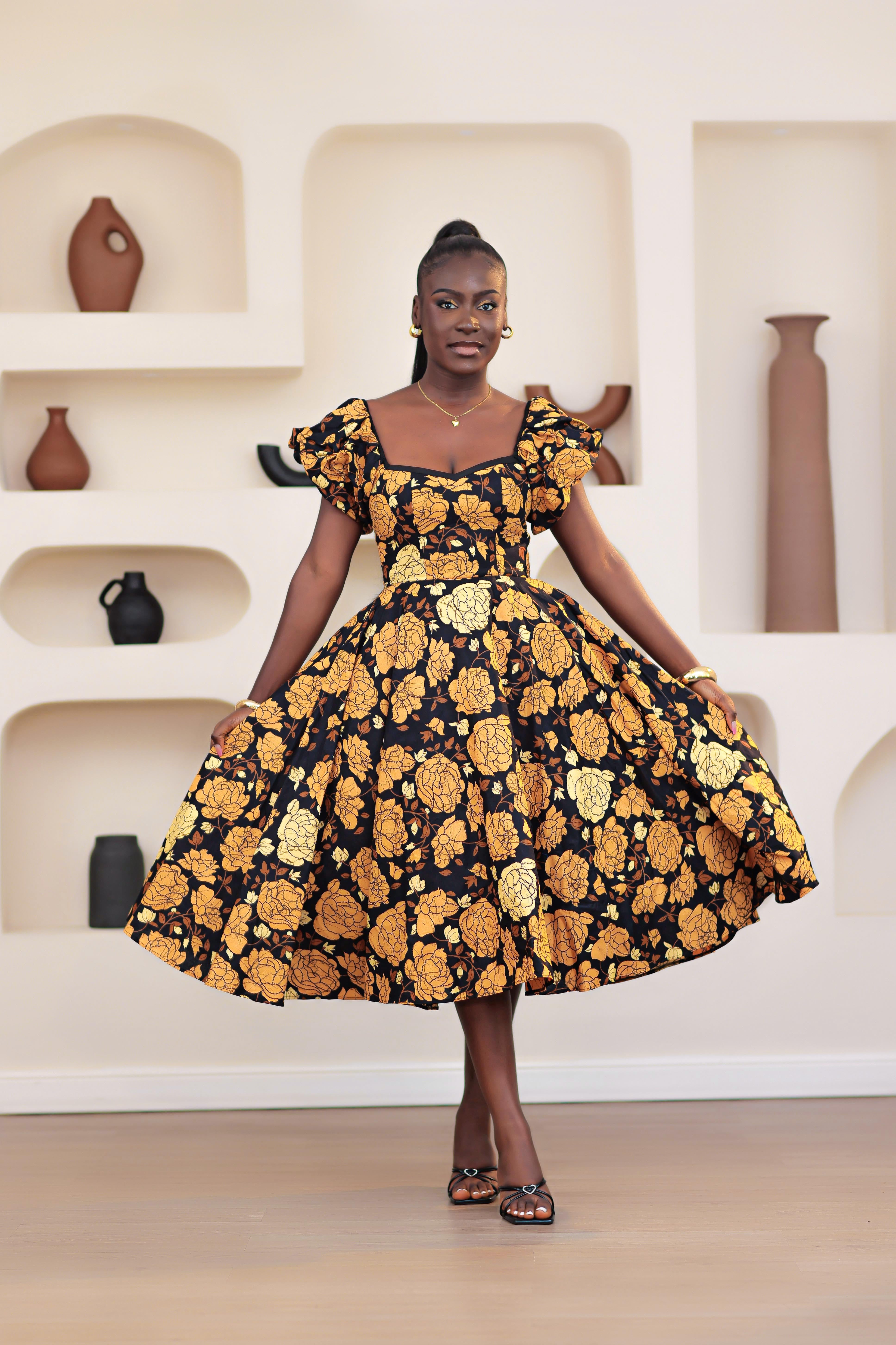 Woman wearing an African print Ankara gold, brown, black, yellow short sleeve floral dress in a room with shelves and vases.