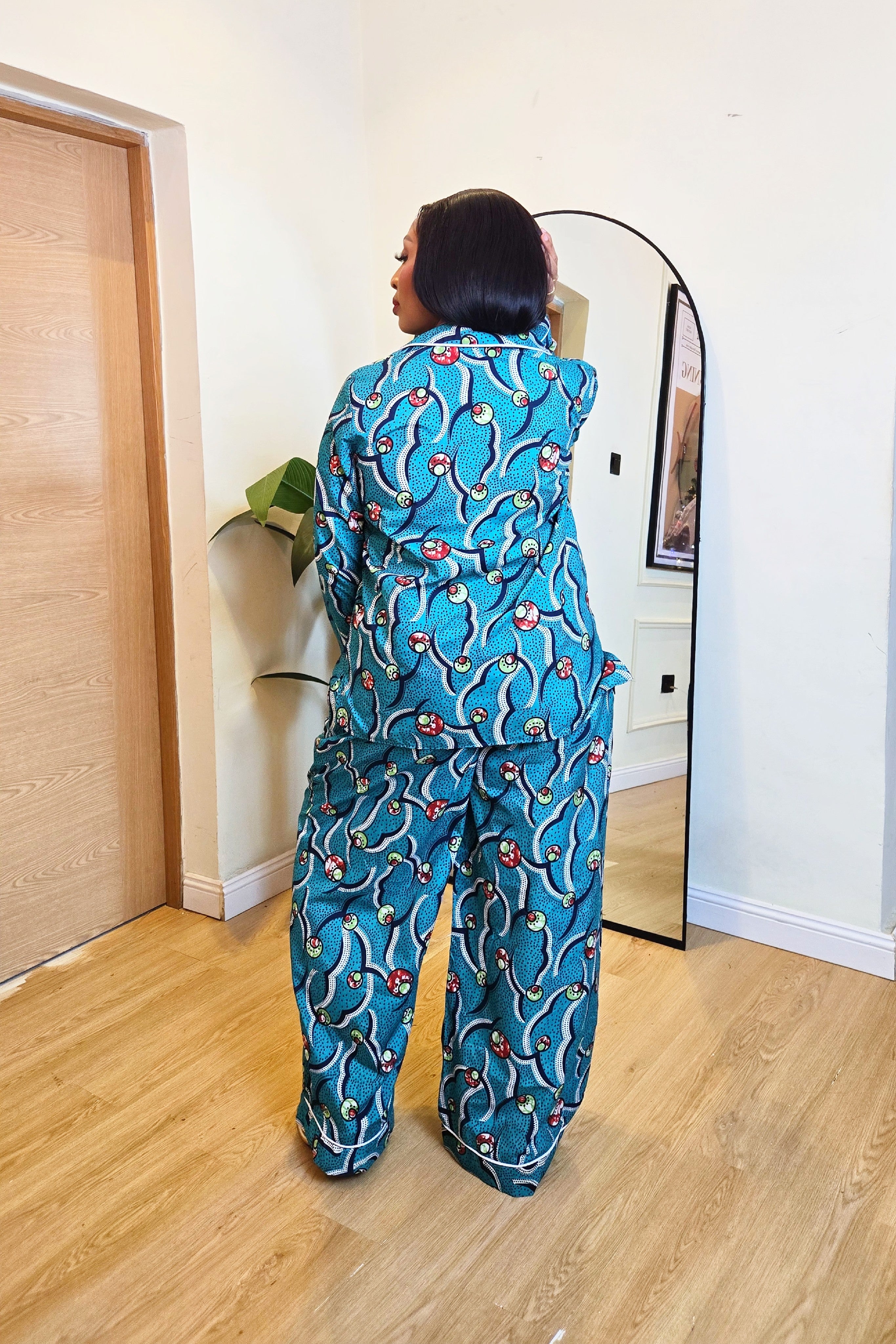 Woman in a colorful green teal African print pajamas shirt and pants set standing in front of a mirror in a room with wooden flooring.