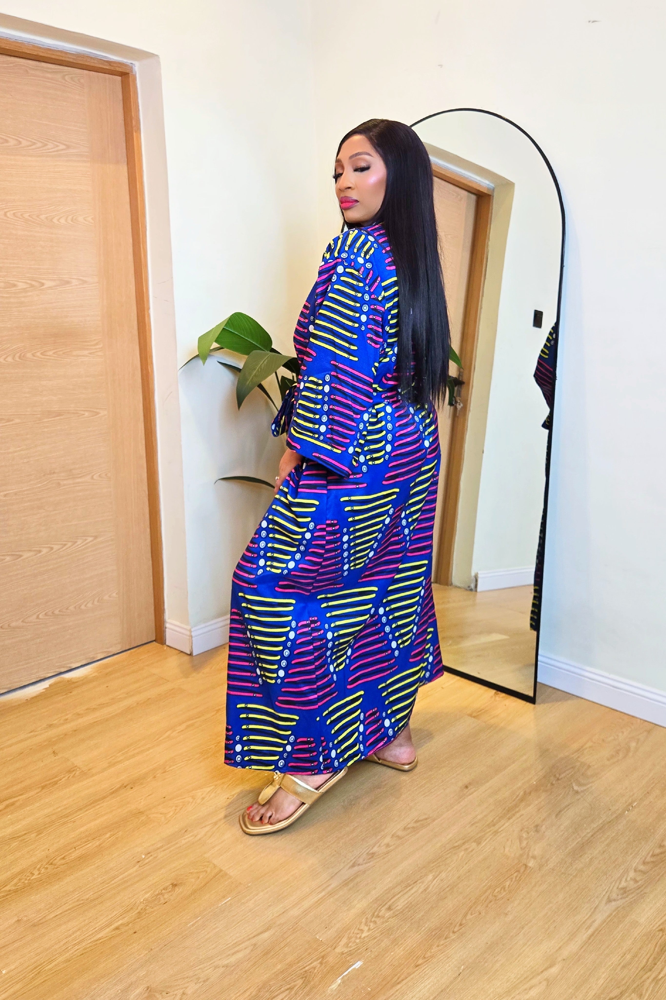 Woman in a colorful African print robe dress standing in front of a mirror in a room with wooden flooring.