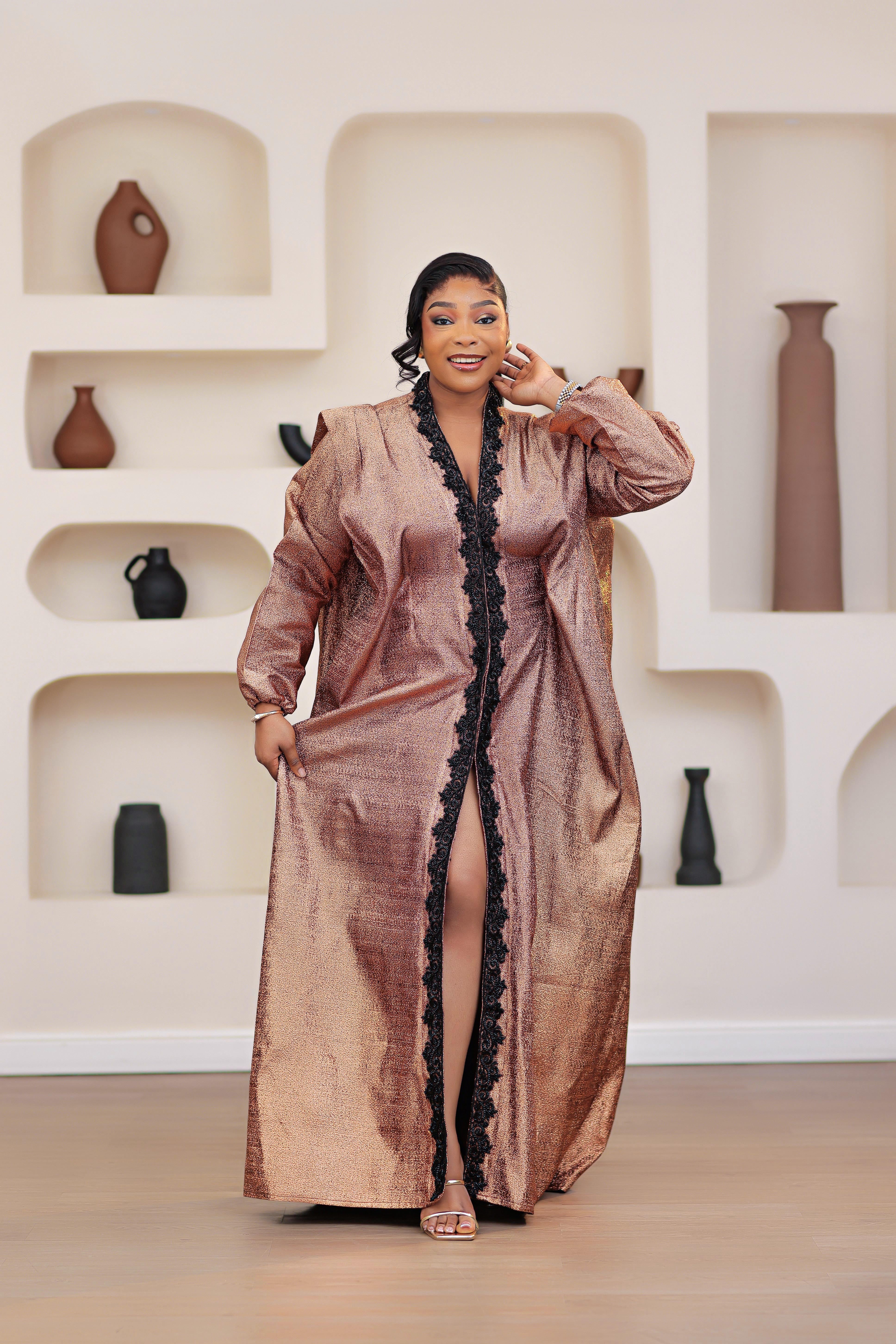 Woman in a long, African brocade boubou dress with embellished black stone trimmings standing in a room with shelves displaying vases.