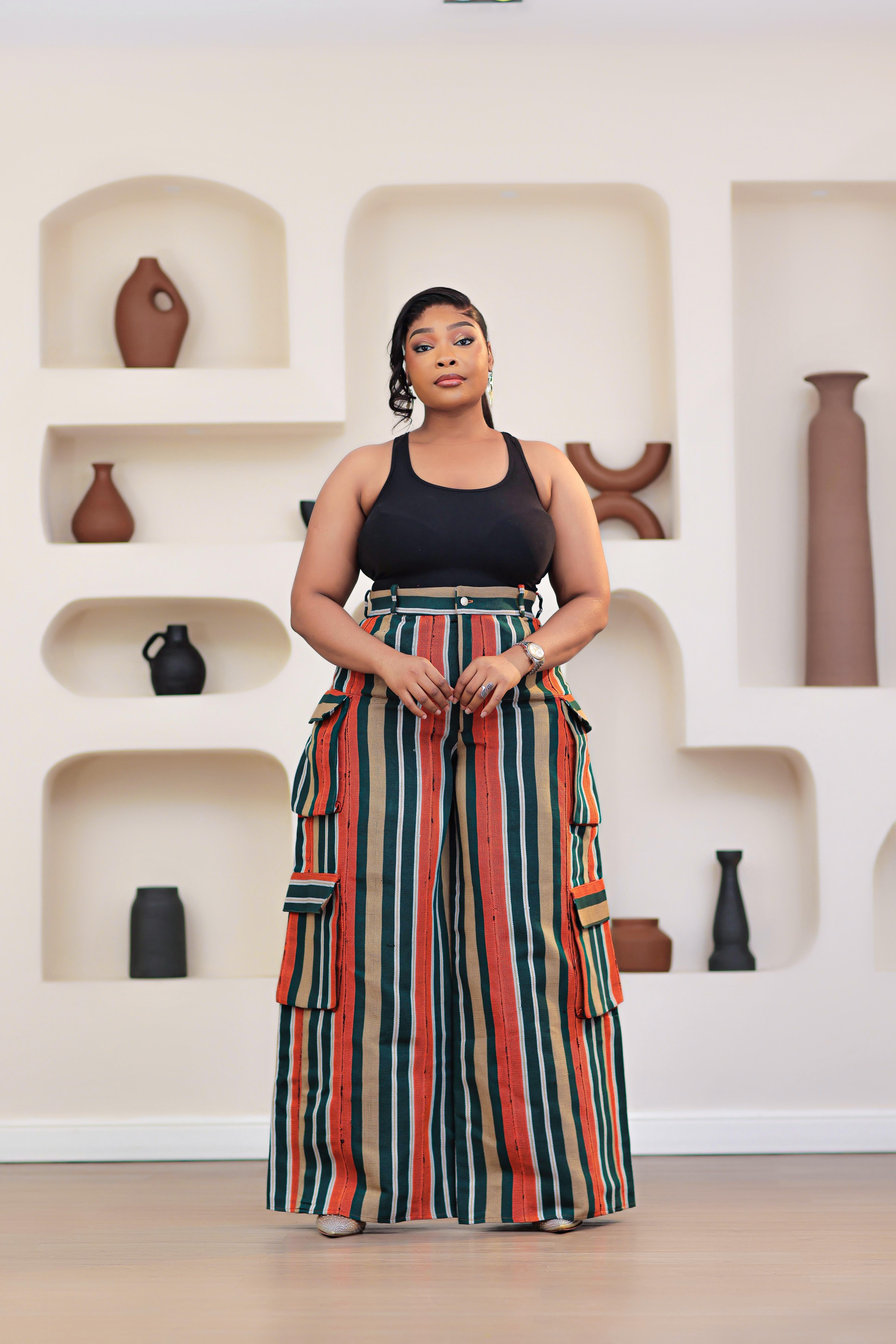 Woman wearing a black top and colorful striped African Aso-oke cargo pants by NIKI KAY standing in front of a white wall with shelves displaying vases.