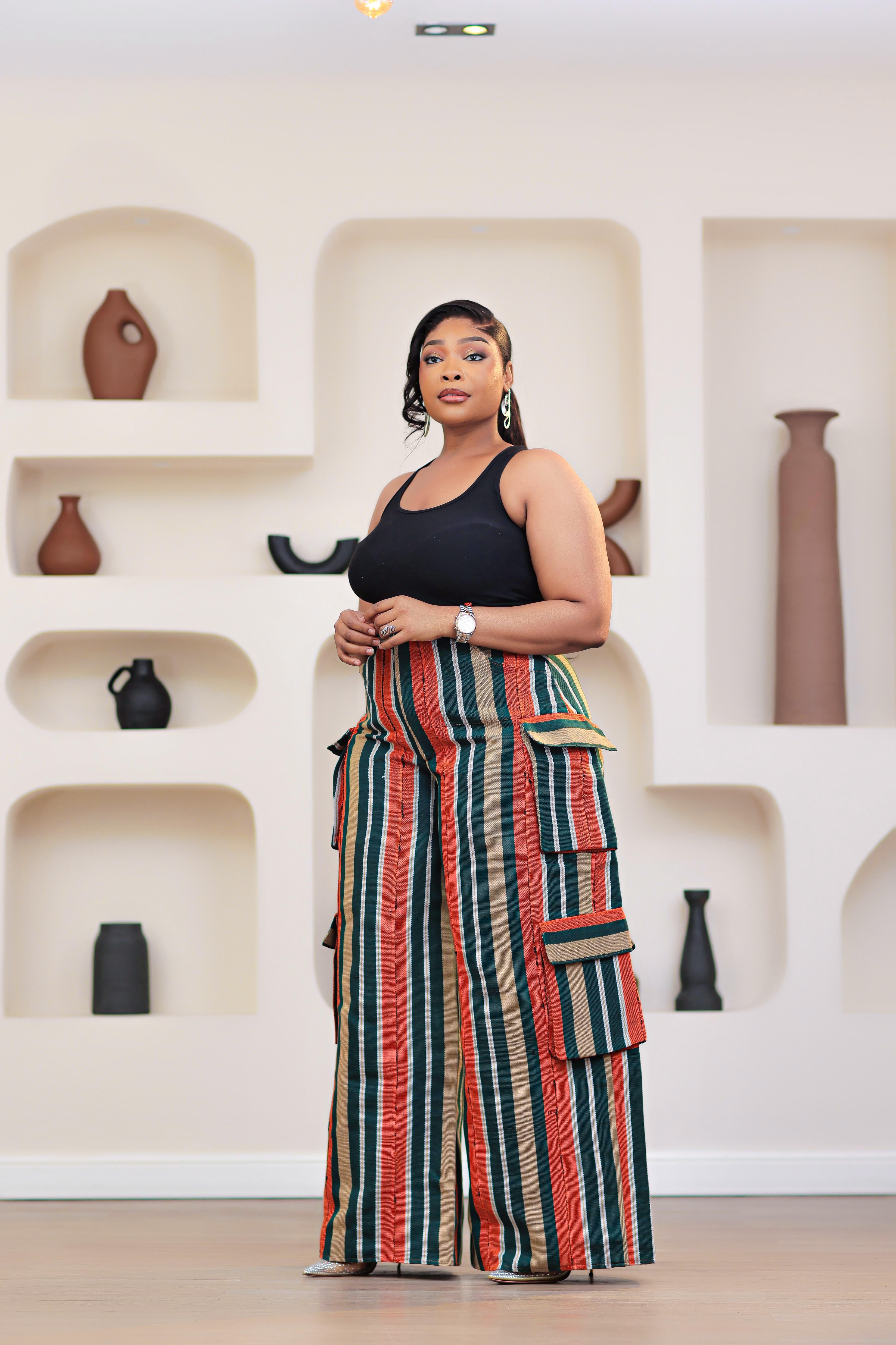 Woman wearing a black top and colorful striped African Aso-oke cargo pants by NIKI KAY standing in front of a white wall with shelves displaying vases.