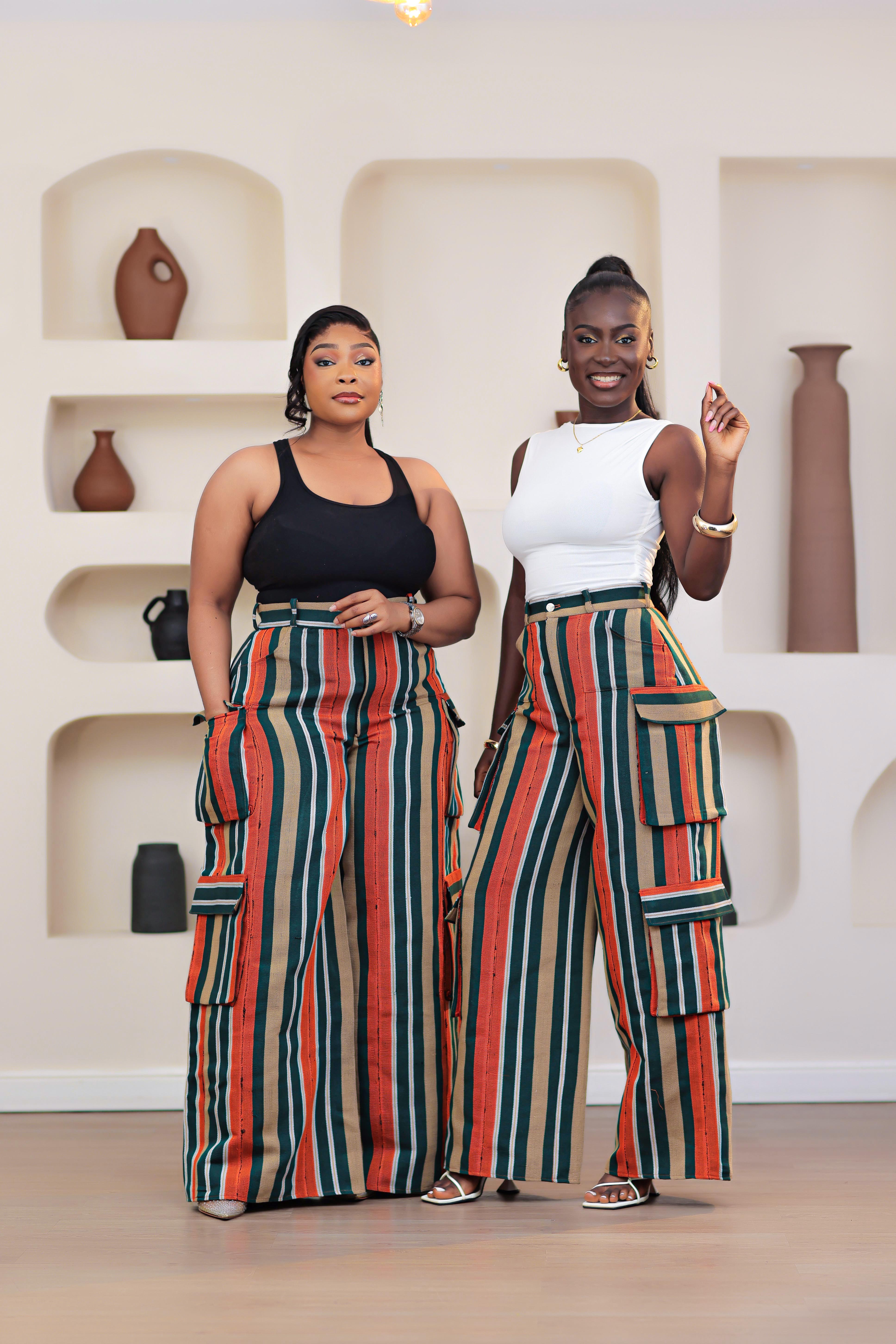 Woman wearing a black top and colorful striped African Aso-oke cargo pants by NIKI KAY standing in front of a white wall with shelves displaying vases.