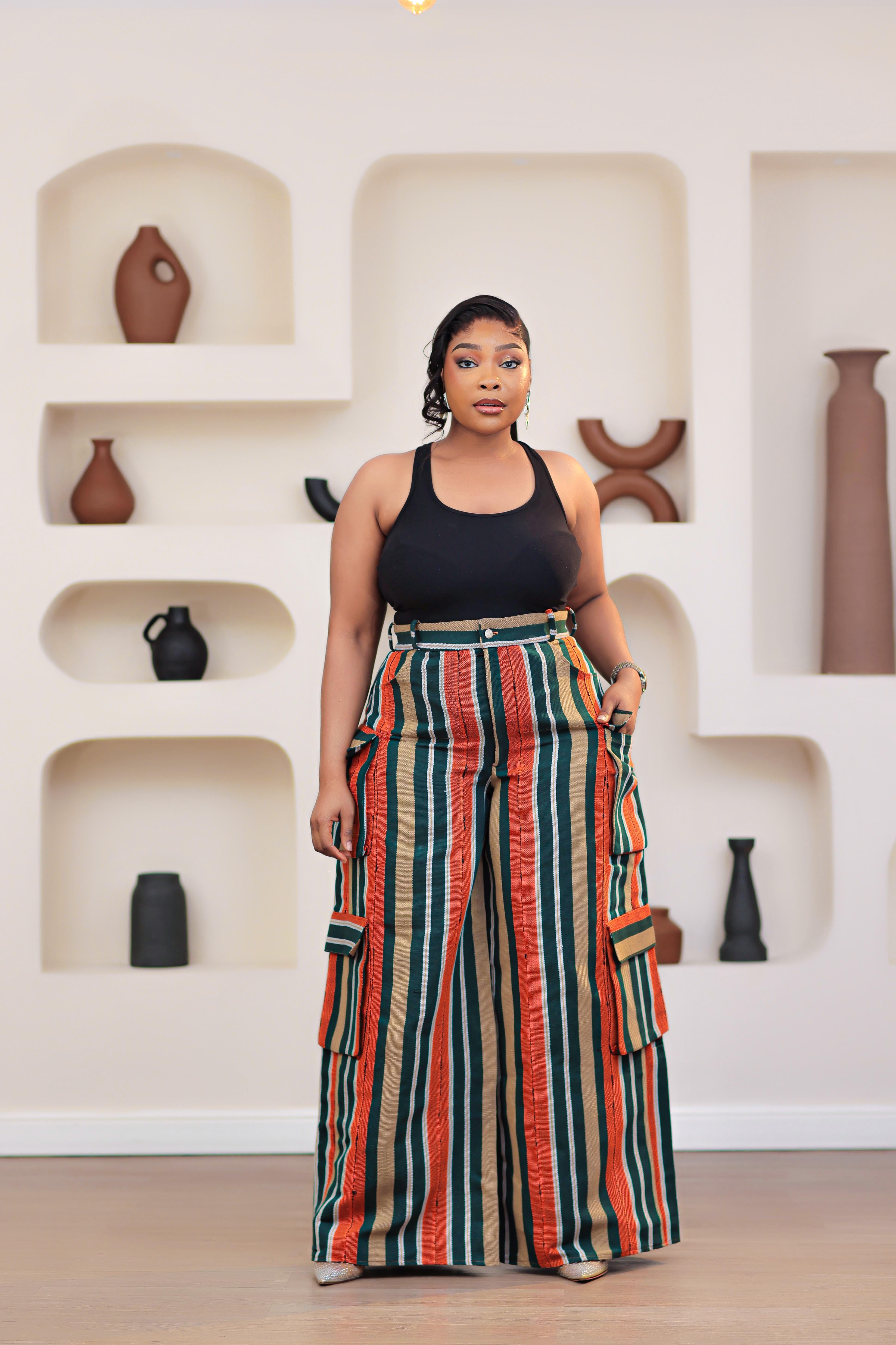 Woman wearing a black top and colorful striped African aso-oke cargo pants in a room with shelves and vases.
