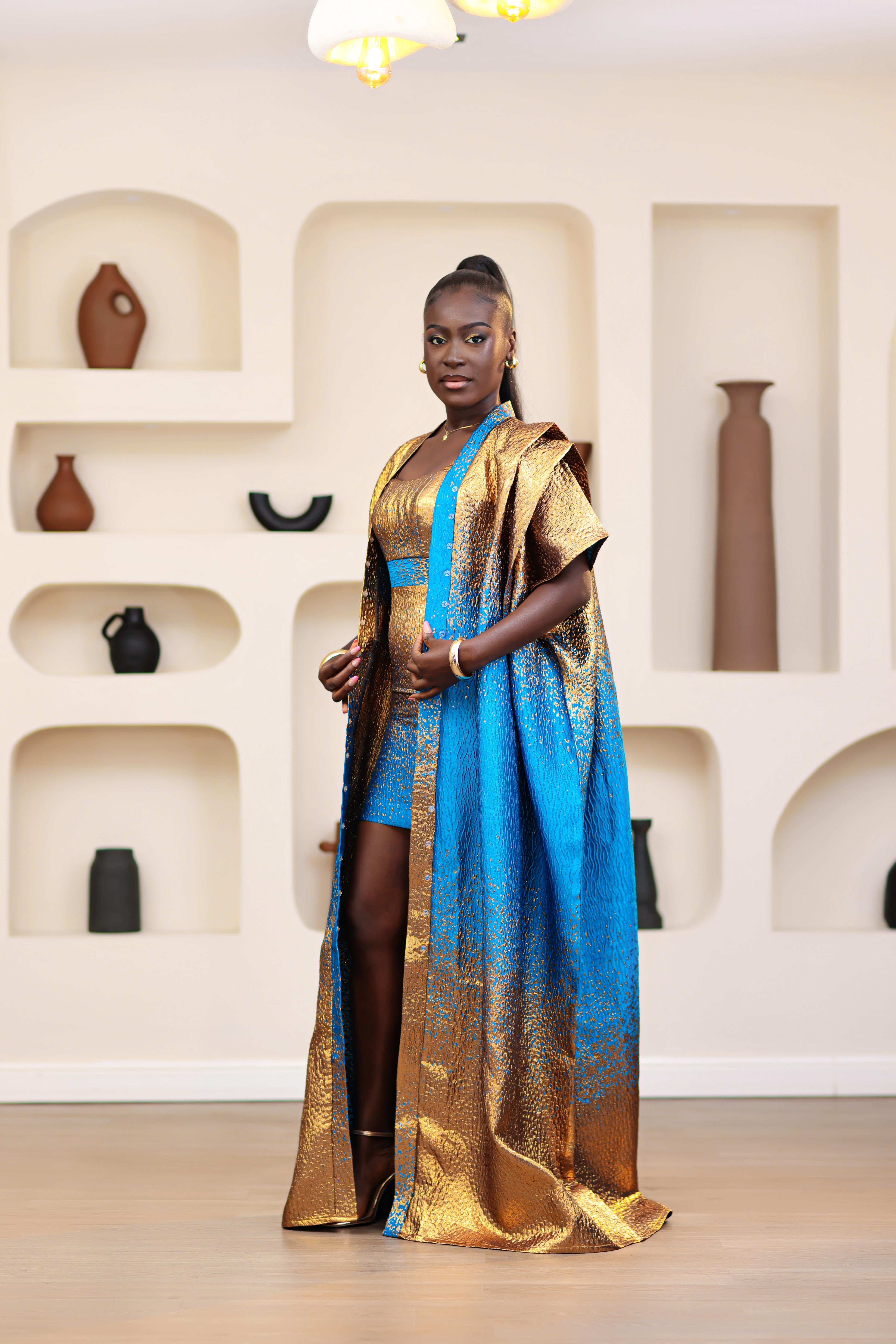 Woman in a blue and gold African traditional Brocade boubou and mini dress outfit set standing in a room with shelves displaying vases.