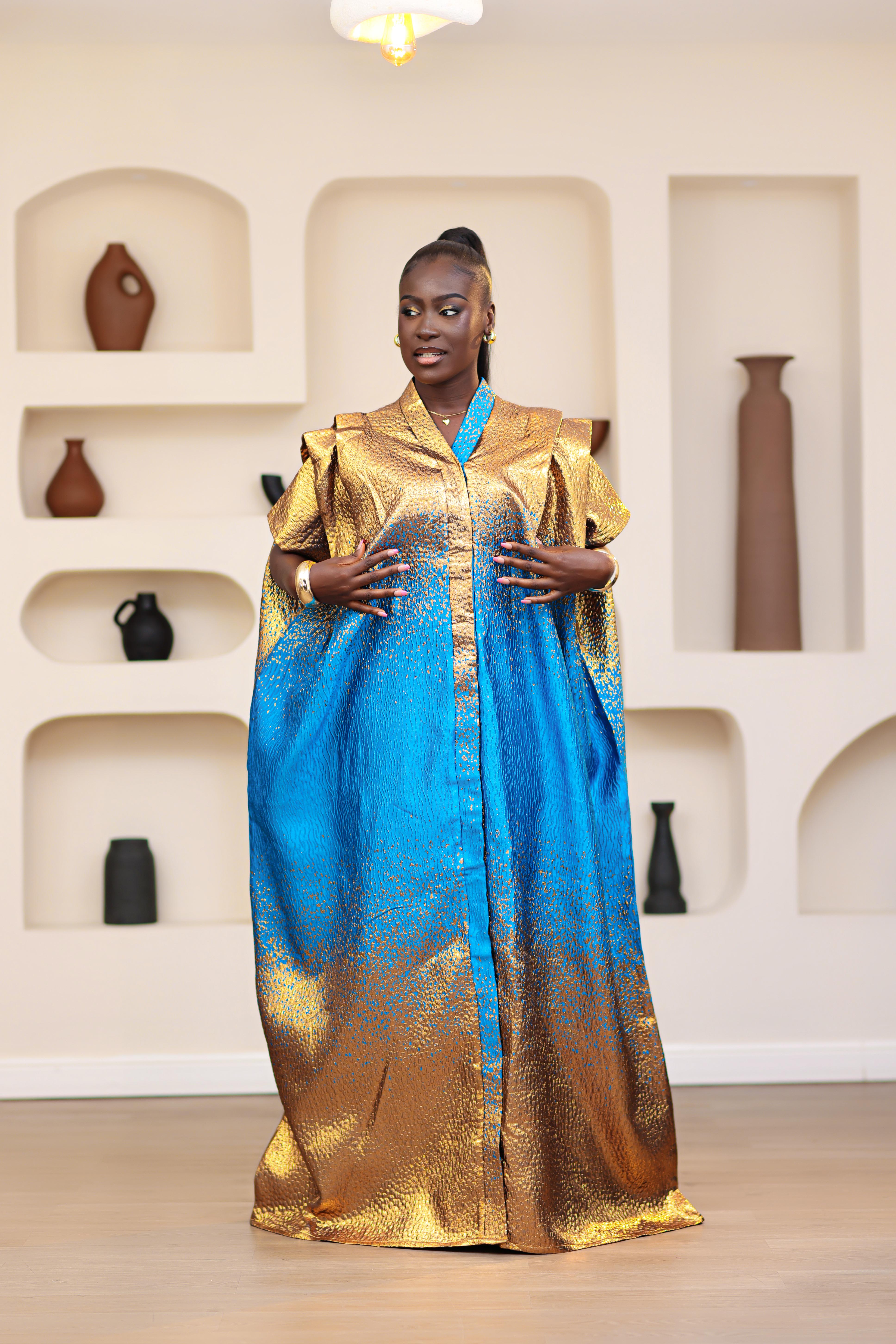 Woman in a blue and gold African traditional Brocade boubou and mini dress outfit set standing in a room with shelves displaying vases.