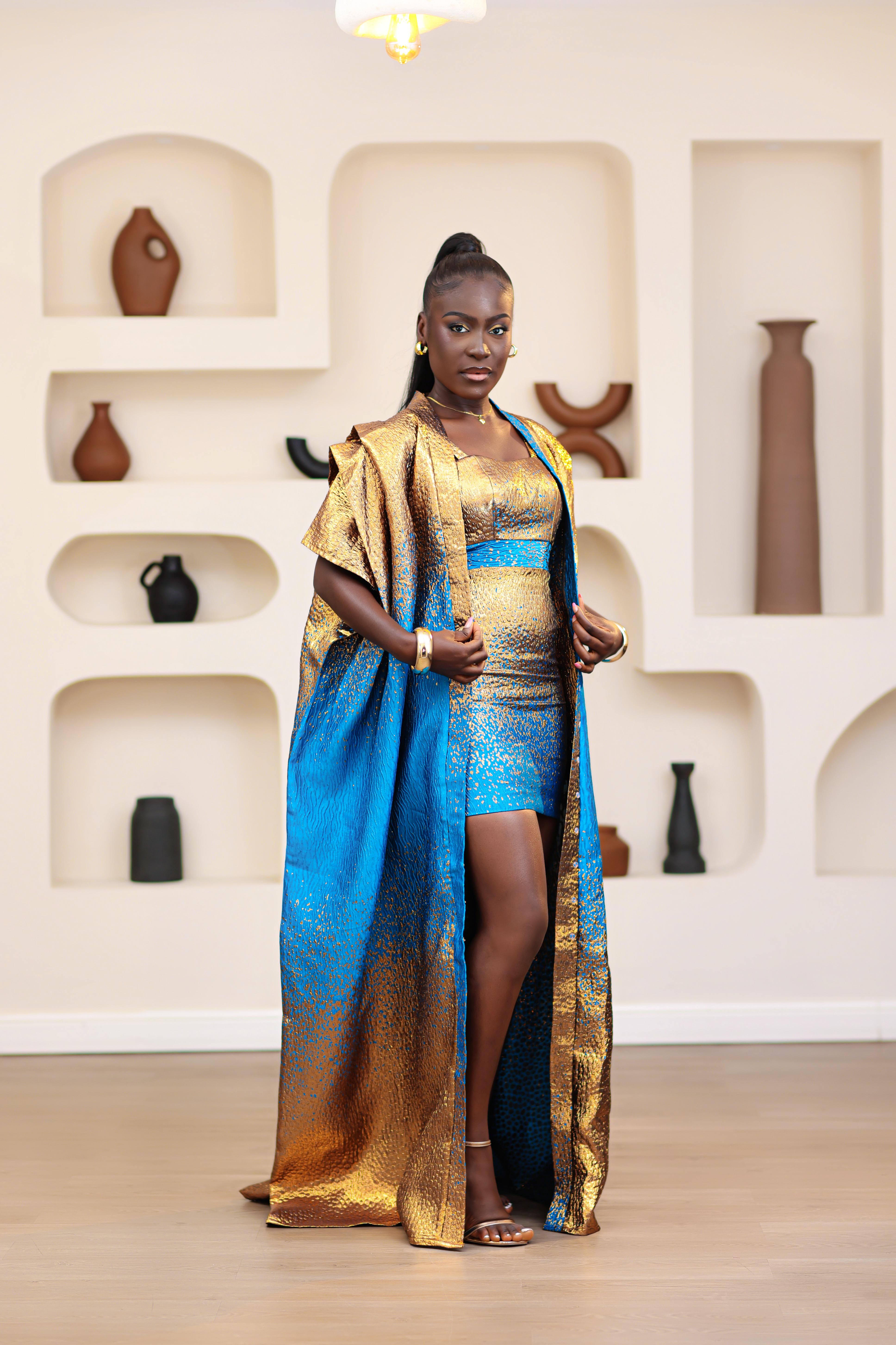 Woman in a blue and gold African traditional Brocade boubou and mini dress outfit set standing in a room with shelves displaying vases.