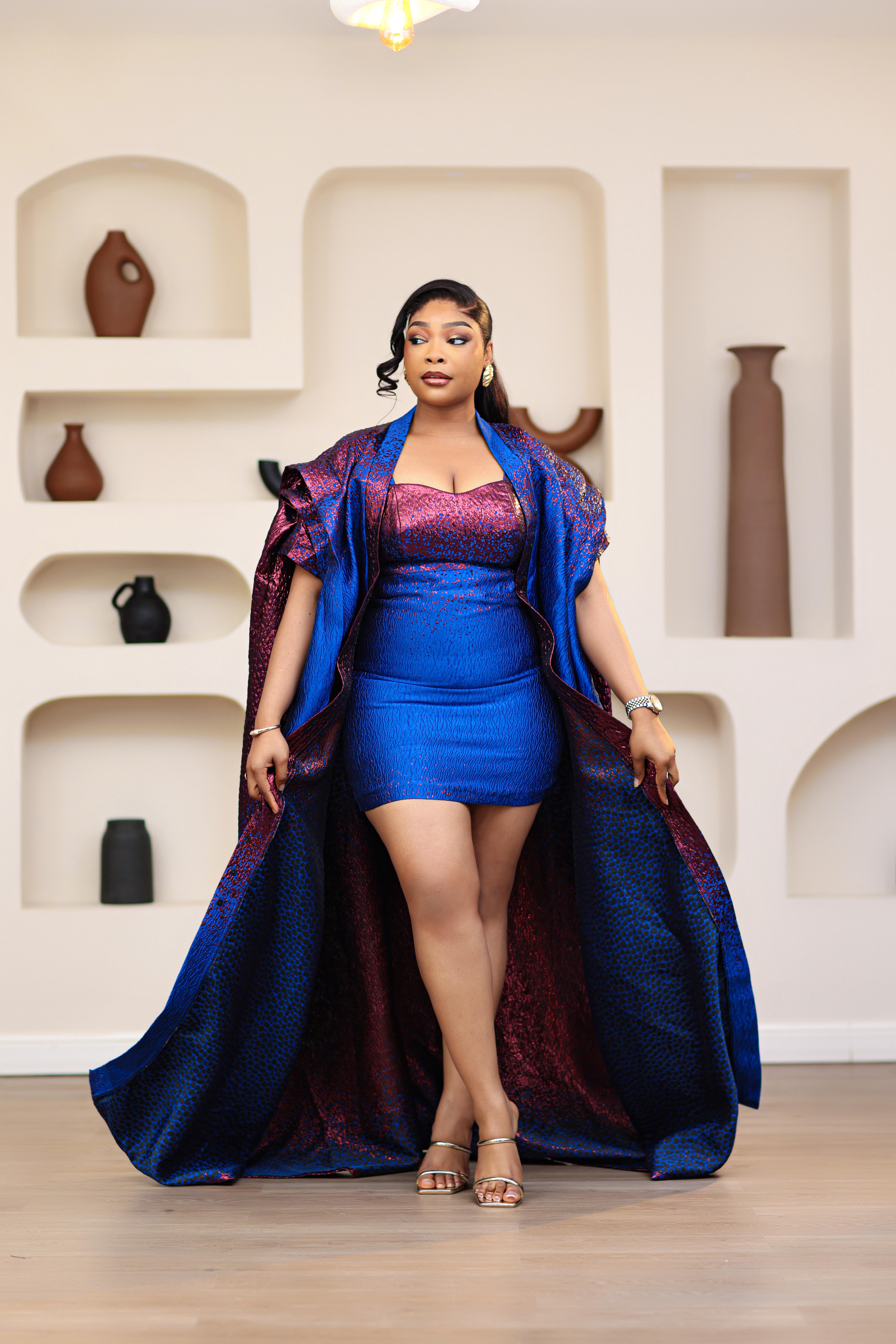 Woman in an African brocade blue and pink mini dress with a matching long boubou kimono standing in a room with shelves displaying vases.
