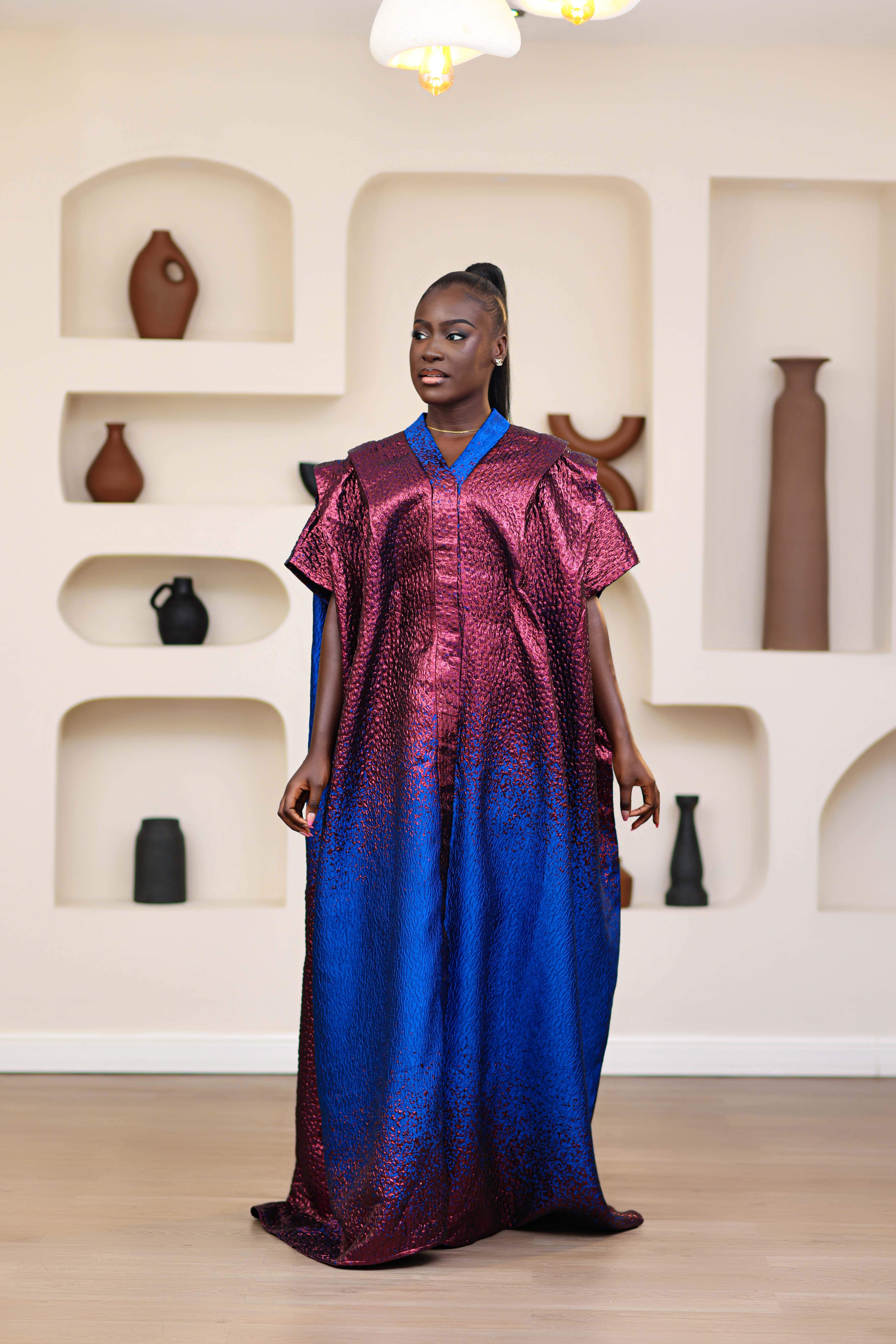 Woman in an African brocade blue and pink mini dress with a matching long boubou kimono standing in a room with shelves displaying vases.