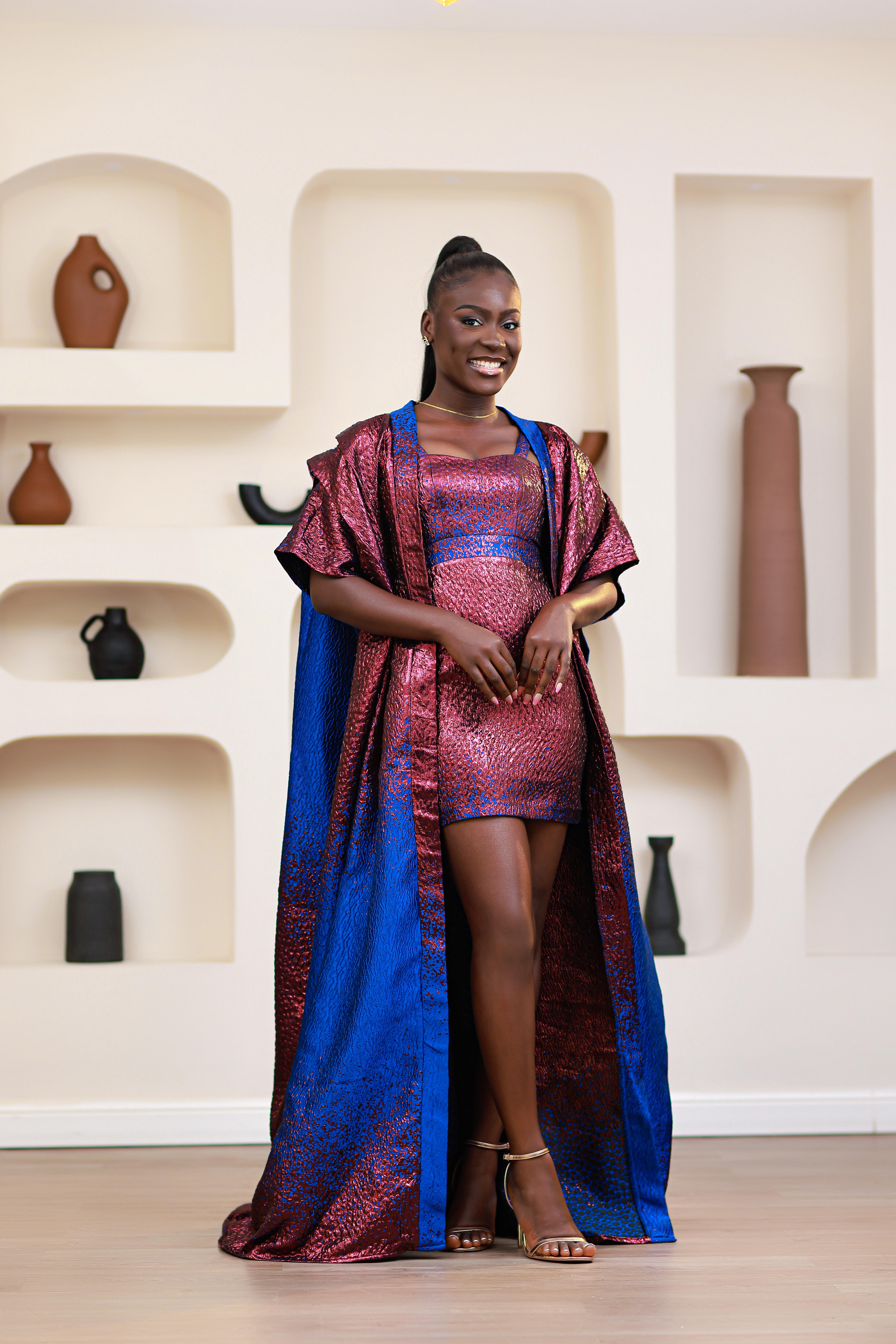 Woman in an African brocade blue and pink mini dress with a matching long boubou kimono standing in a room with shelves displaying vases.