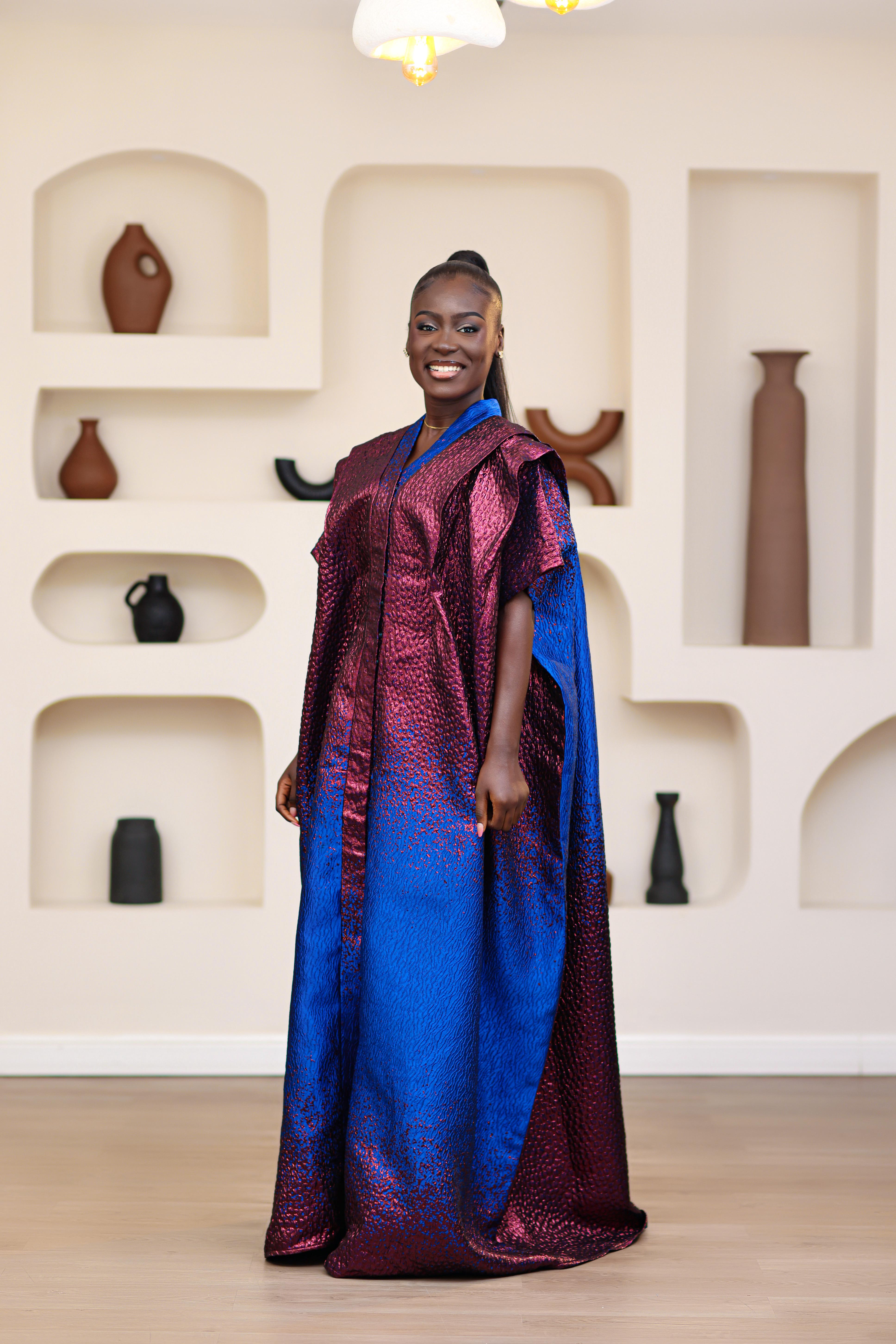 Woman in an African brocade blue and pink mini dress with a matching long boubou kimono standing in a room with shelves displaying vases.