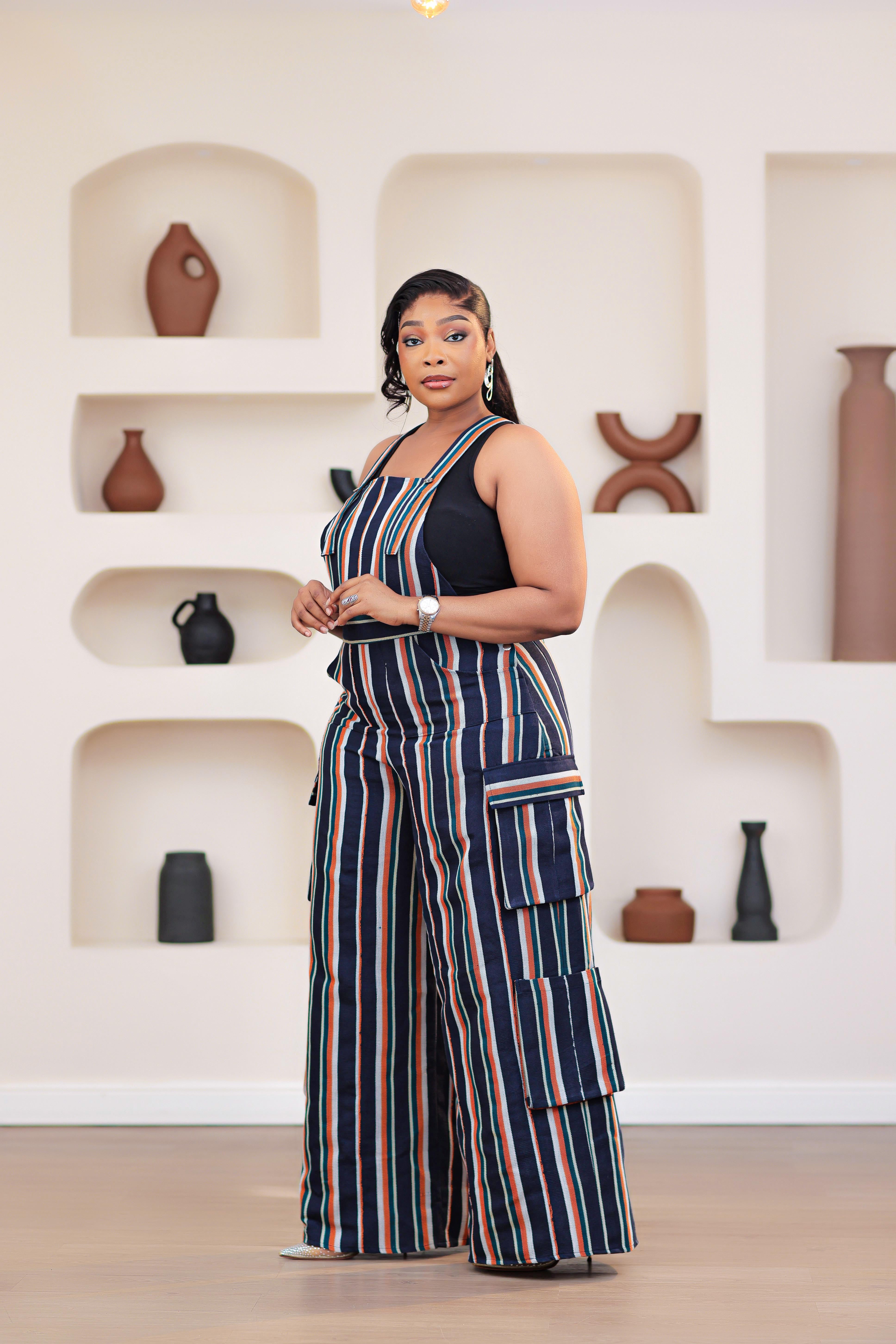 Woman wearing a striped African Aso-oke dungarees jumpsuit standing in front of a decorative wall with shelves.