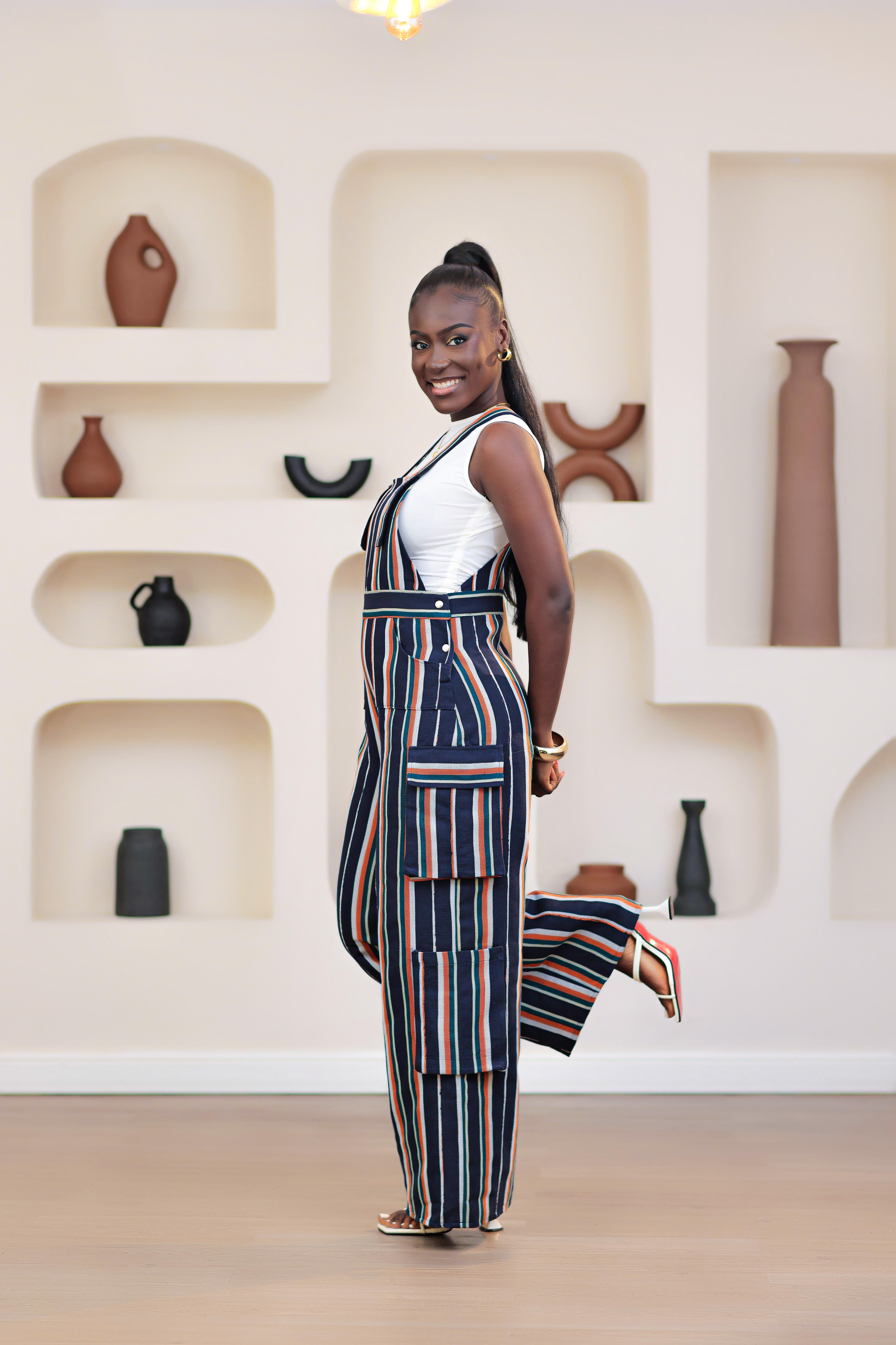 Woman wearing a striped NIKI KAY Aso-oke dungarees jumpsuit standing in a room with shelves and vases. 