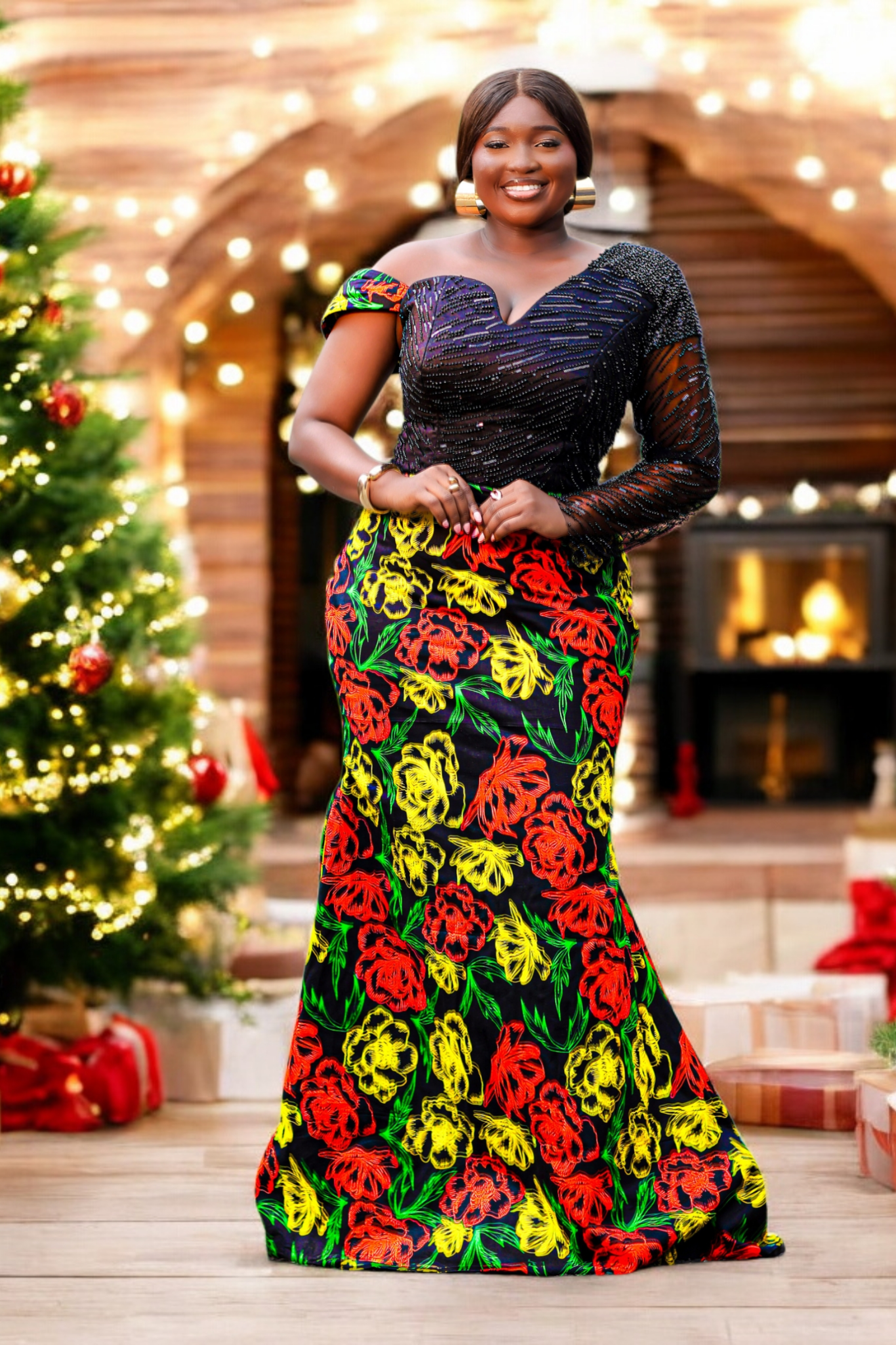 Woman in a colorful African Print dress with embellished hand beaded upper bodice in black standing in front of a decorated Christmas tree and fireplace.