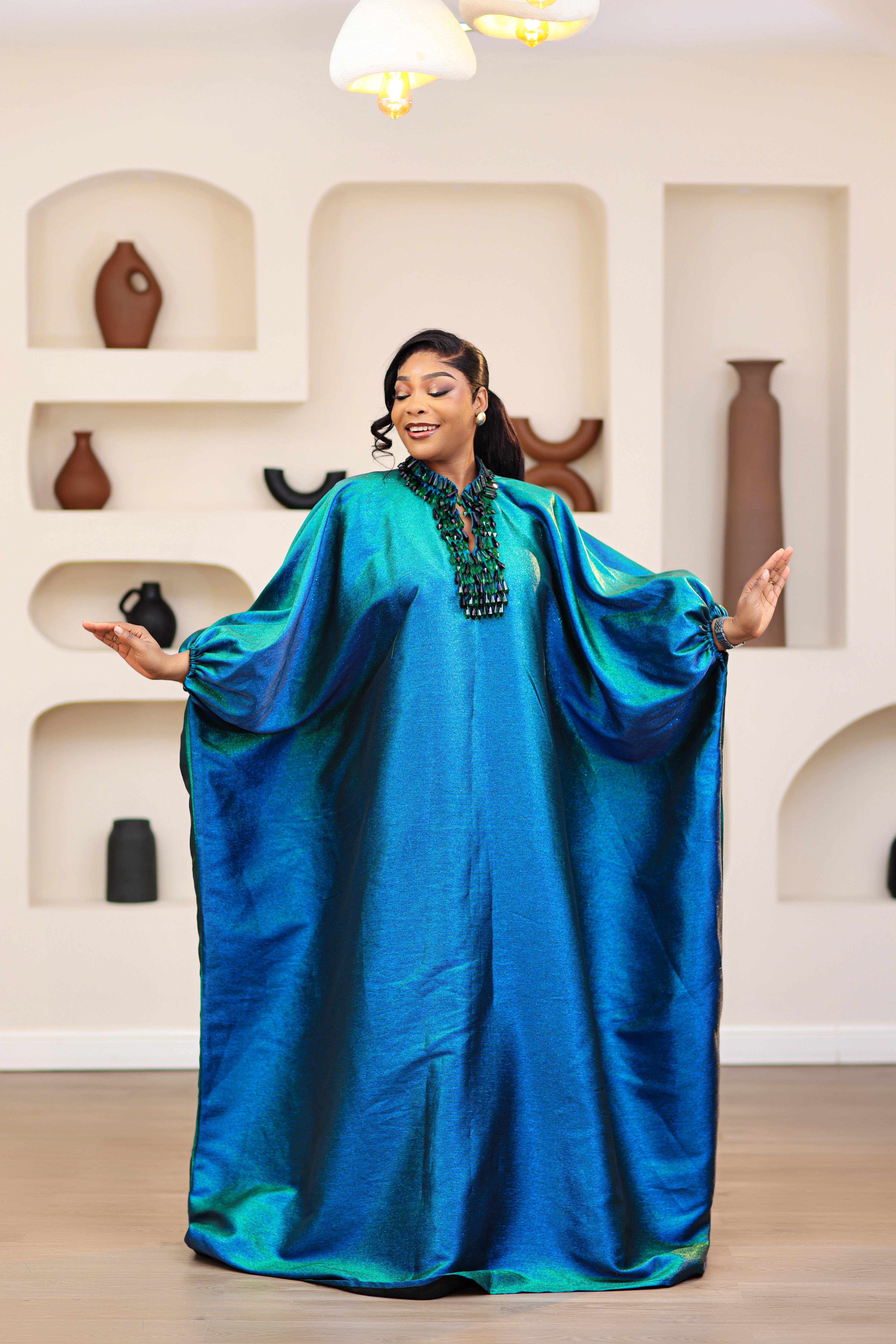 Woman wearing a blue and green NIKI KAY Iridescent brocade boubou dress  with an embellished droplet neckline in a room with decorative vases on shelves.