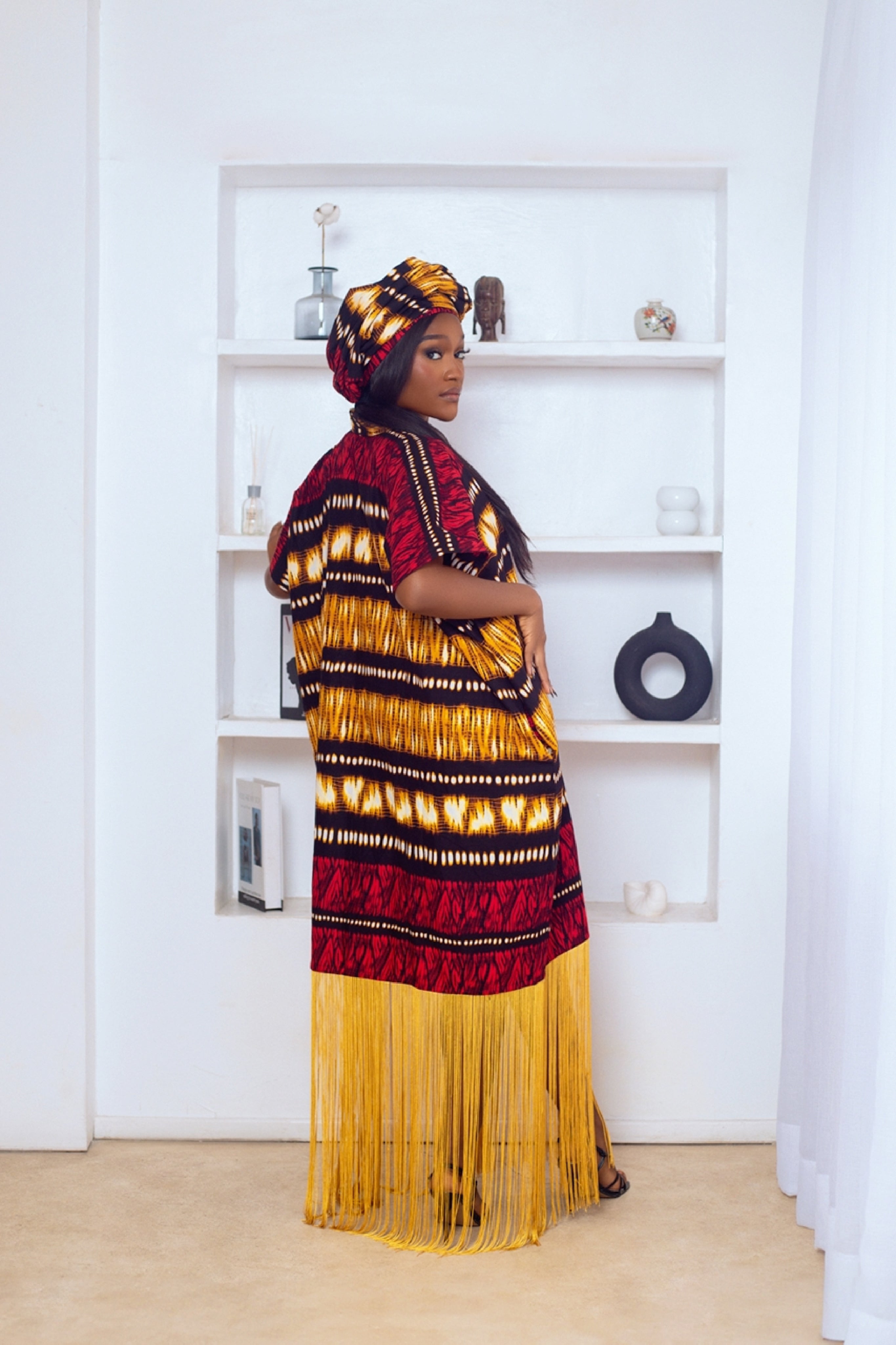 Woman in African Print Ankara Boubou dress outfit with headwrap, fringe and tassle details in red gold brown multicolor