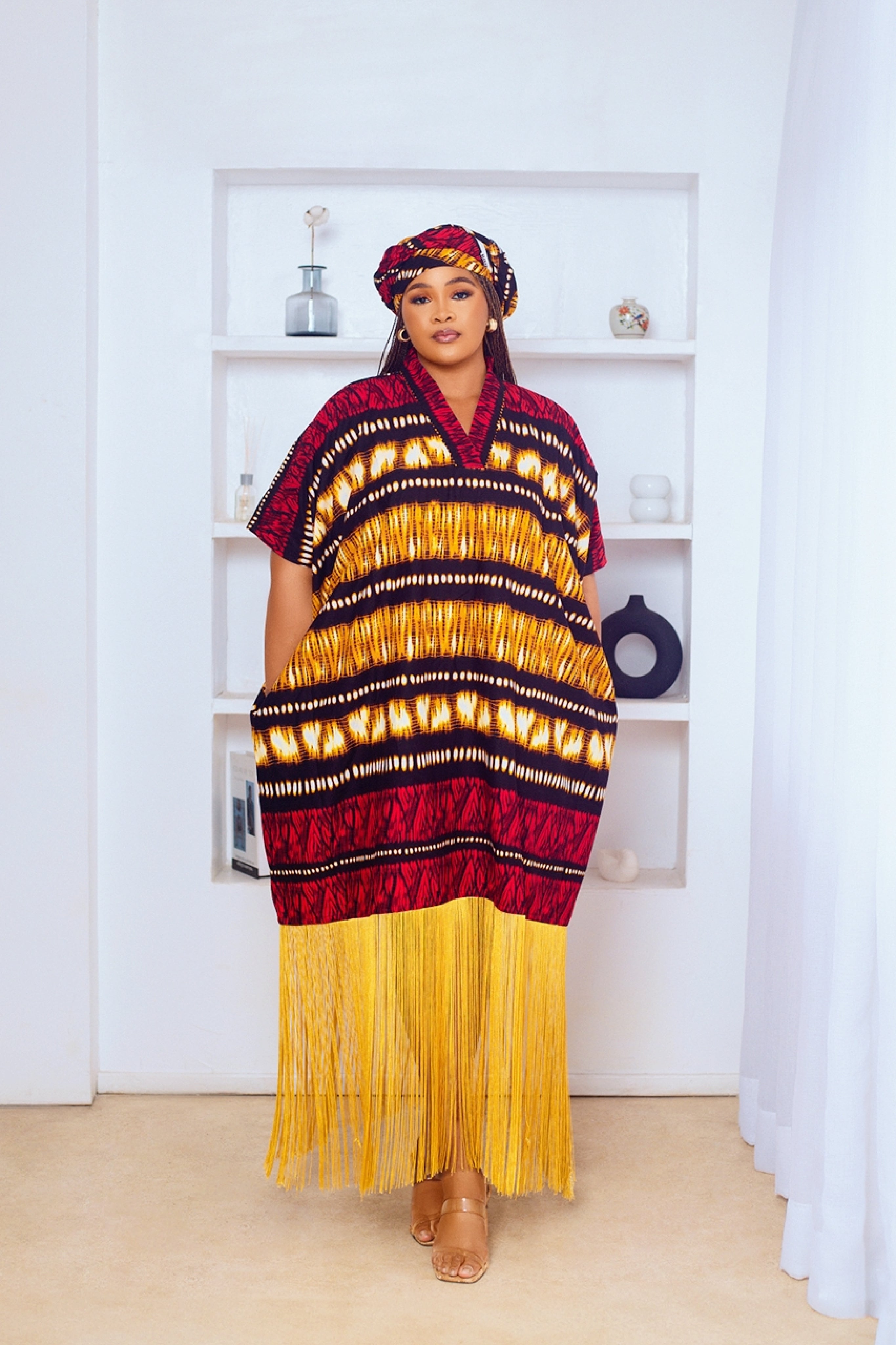Woman in African Print Ankara Boubou dress outfit with headwrap, fringe and tassle details in red gold brown multicolor