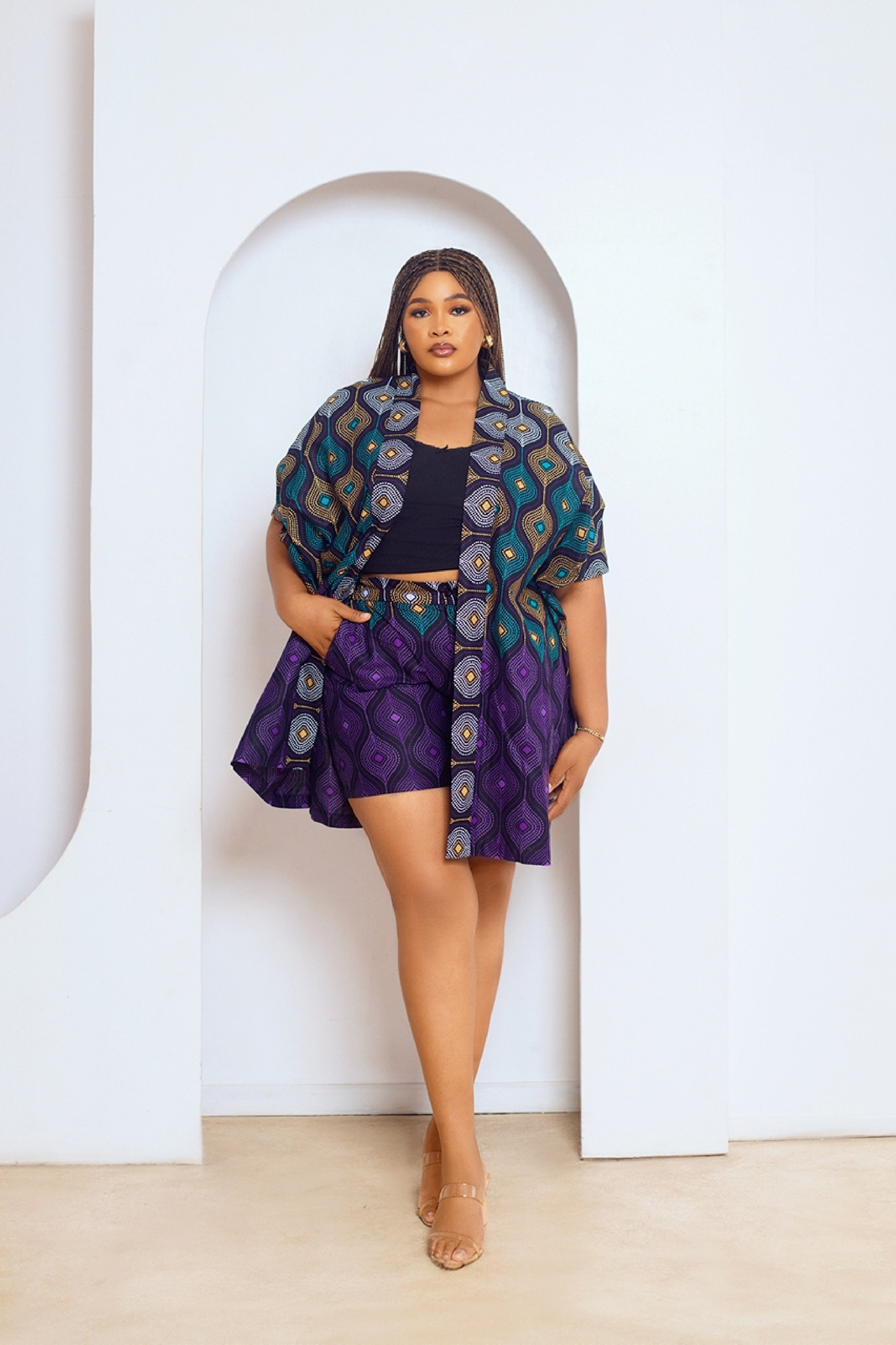A model wearing a multicolored African print kimono and matching shorts with side pockets. The outfit features a mix of purple, green, and other colors in a geometric pattern.