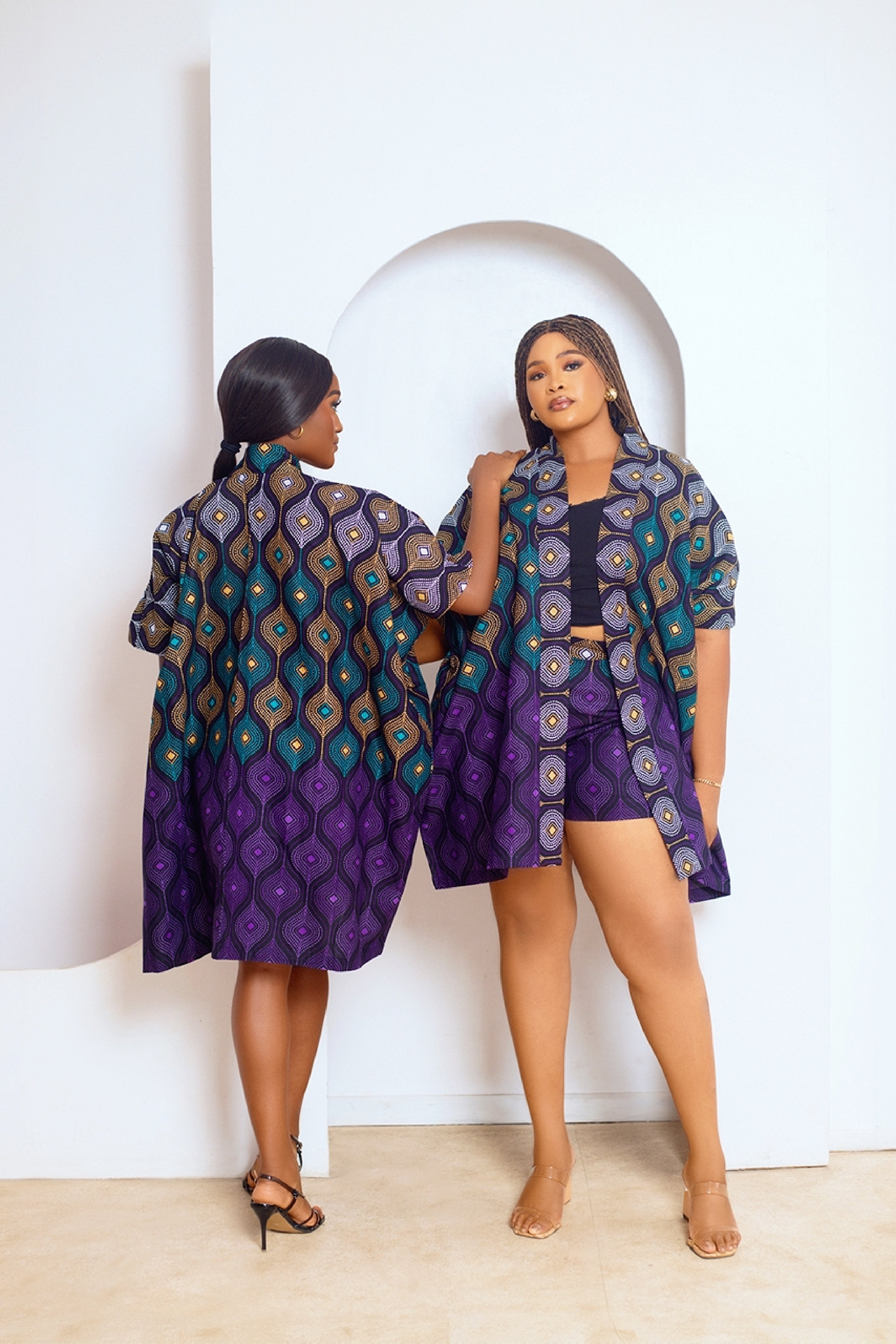 A model wearing a multicolored African print kimono and matching shorts with side pockets. The outfit features a mix of purple, green, and other colors in a geometric pattern.