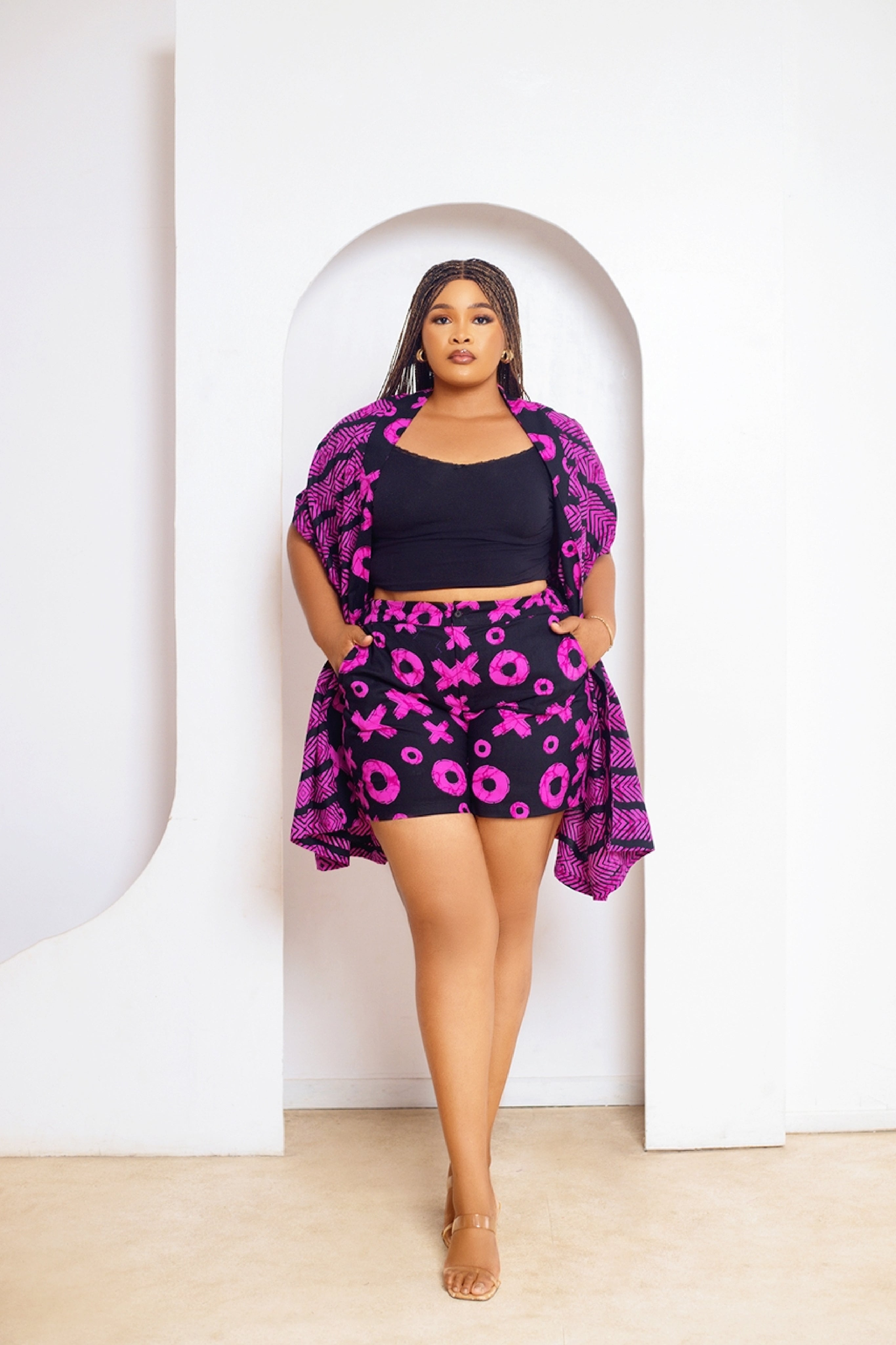 A model wearing a multicolored African print kimono and matching shorts with side pockets. The outfit features a mix of pink and black colors in a geometric pattern.
