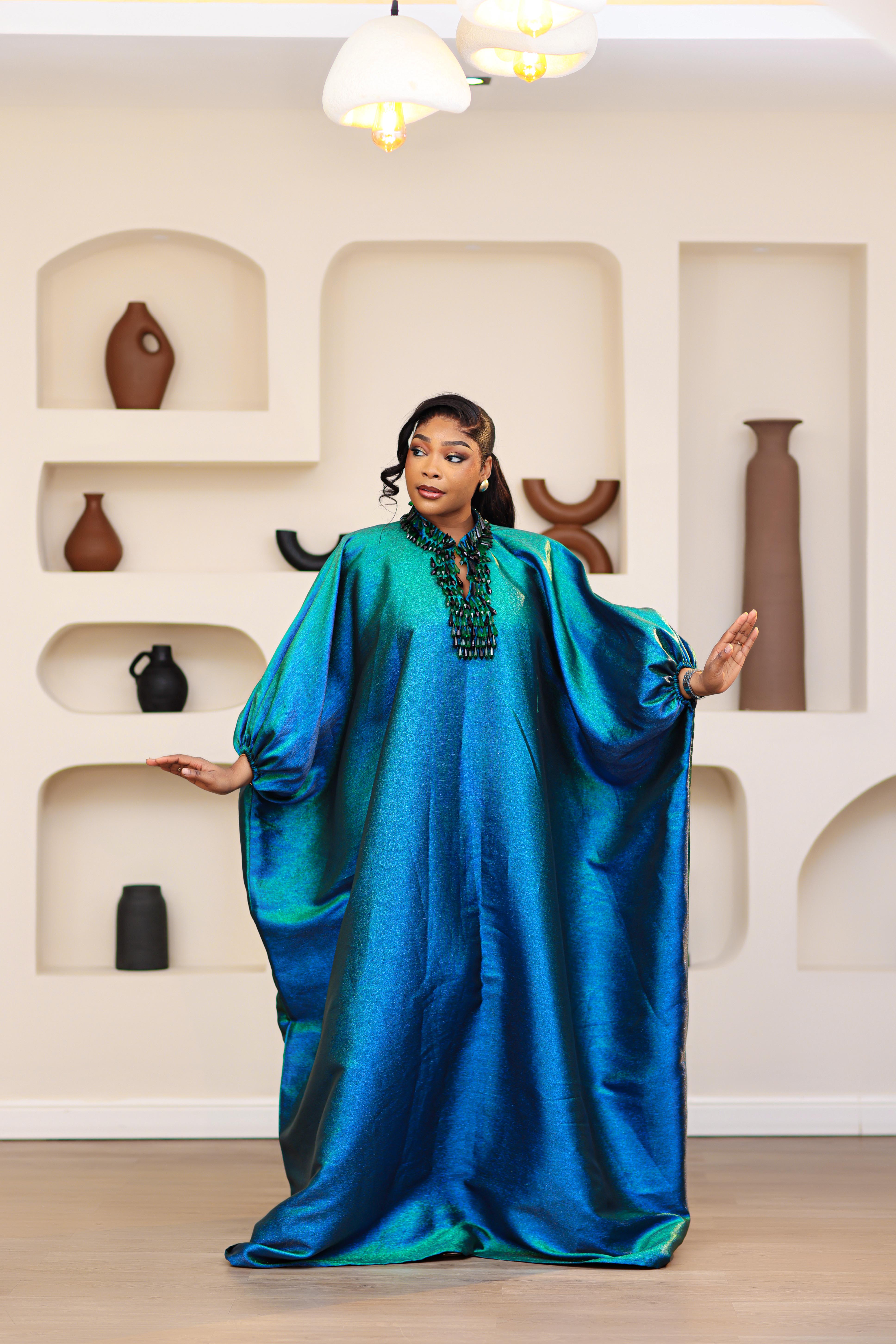 Woman wearing a blue and green NIKI KAY Iridescent brocade boubou dress in a room with decorative vases on shelves.
